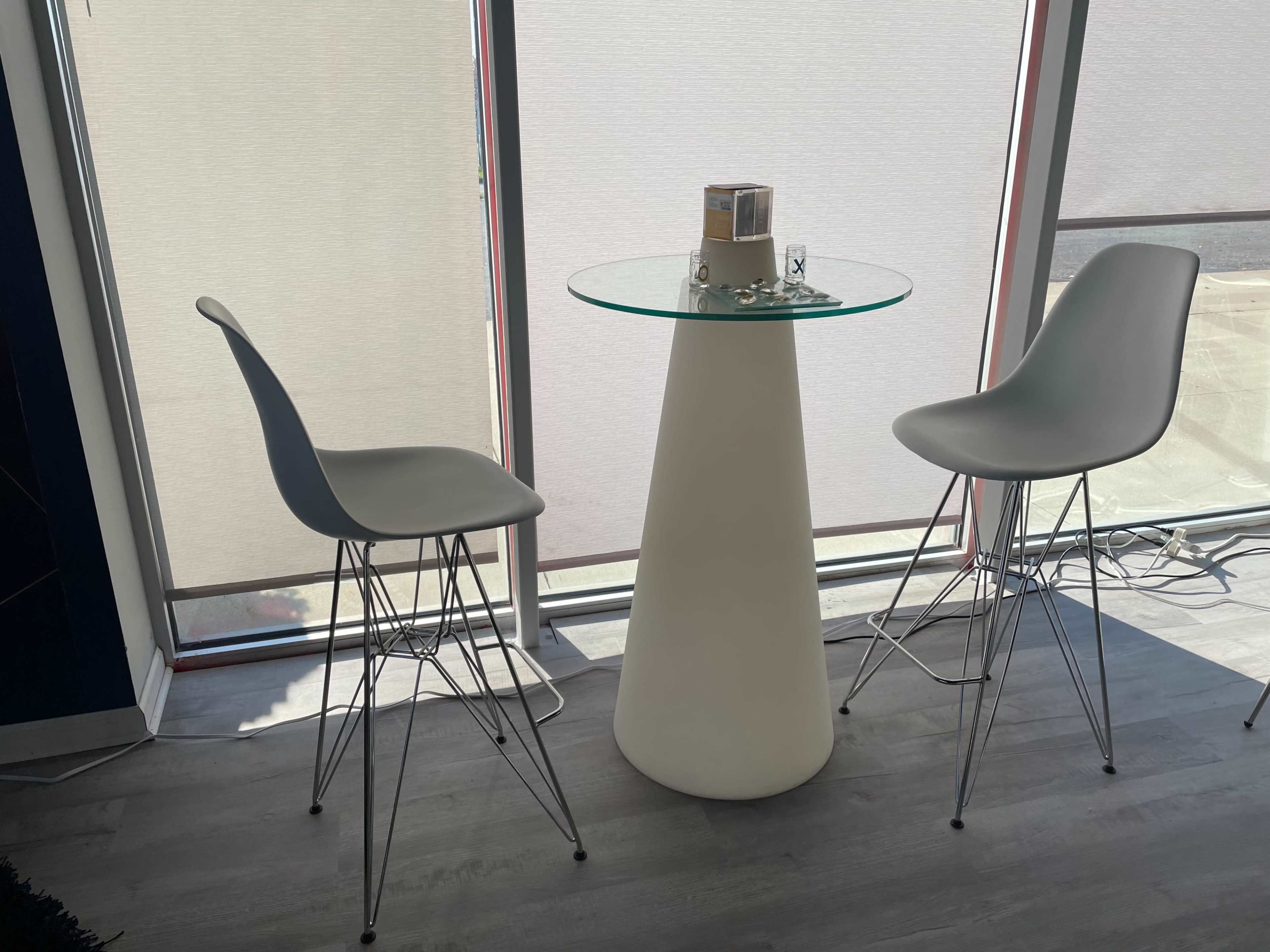 A round glass table stands on a white pedestal between two gray bar stools near a window.