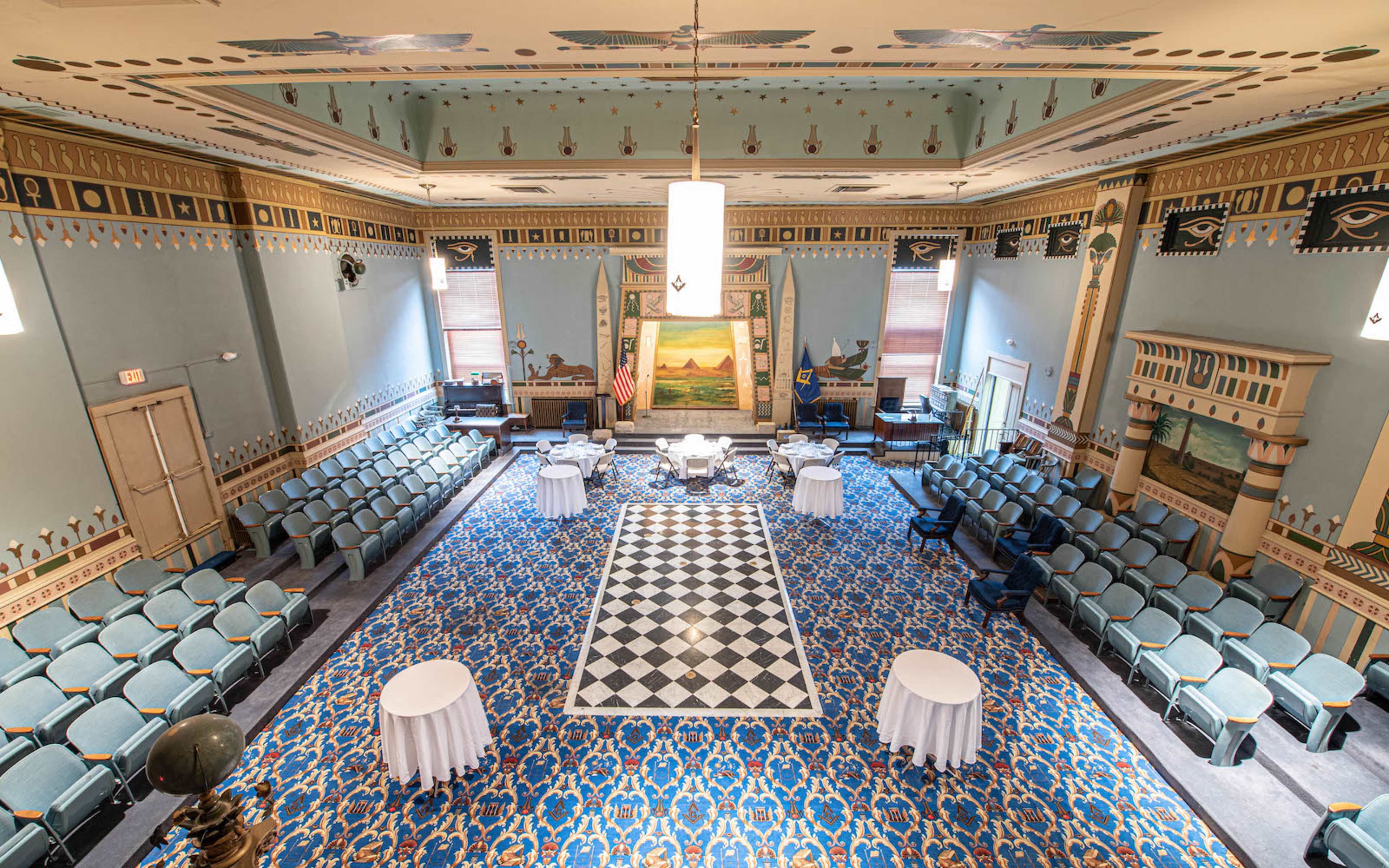 A spacious room set up for an event, featuring a checkered floor, seated rows of chairs, and decorative walls with murals.