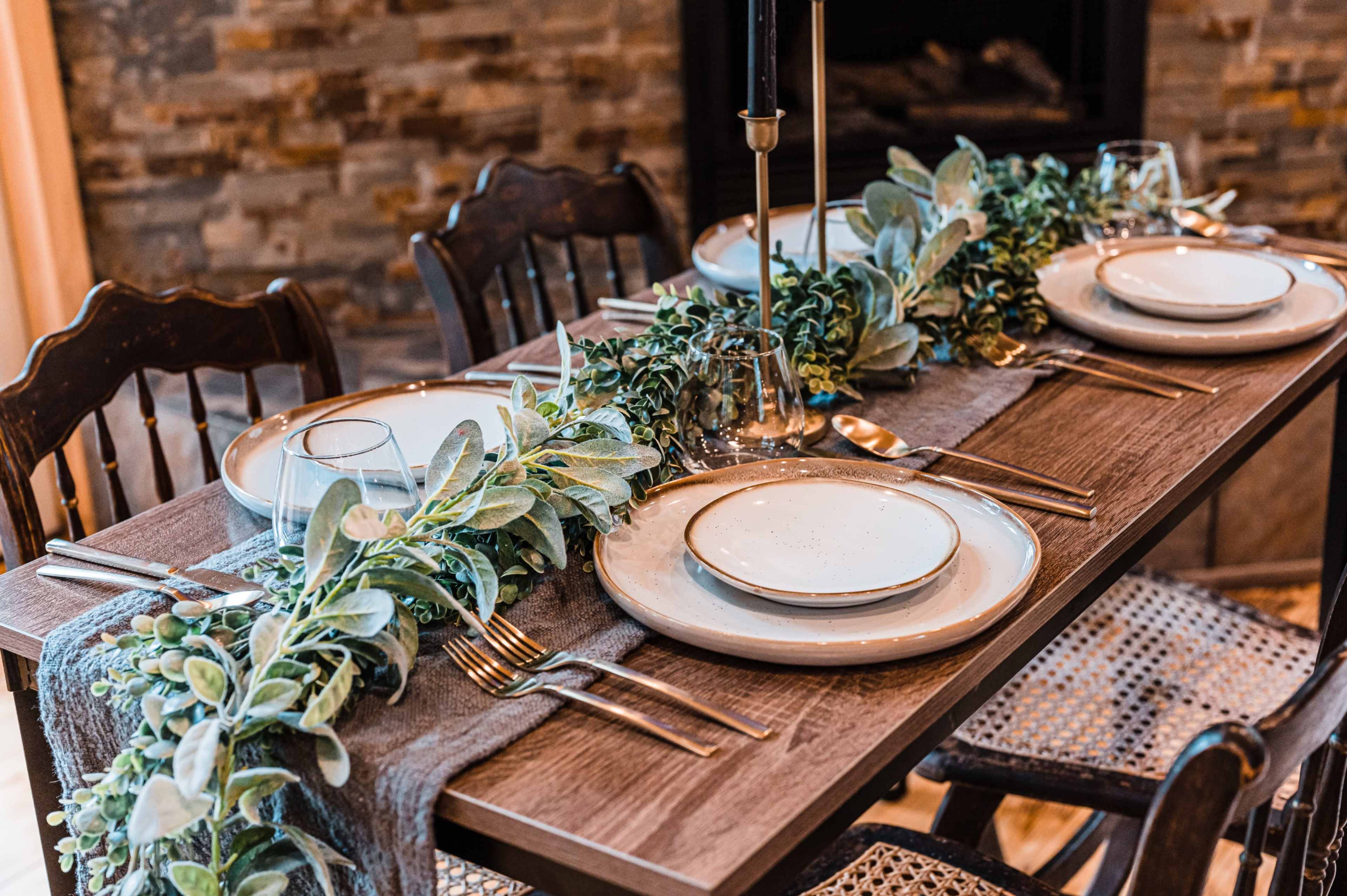 A dining table is set with white plates, silverware, and a greenery centerpiece, surrounded by wooden chairs.