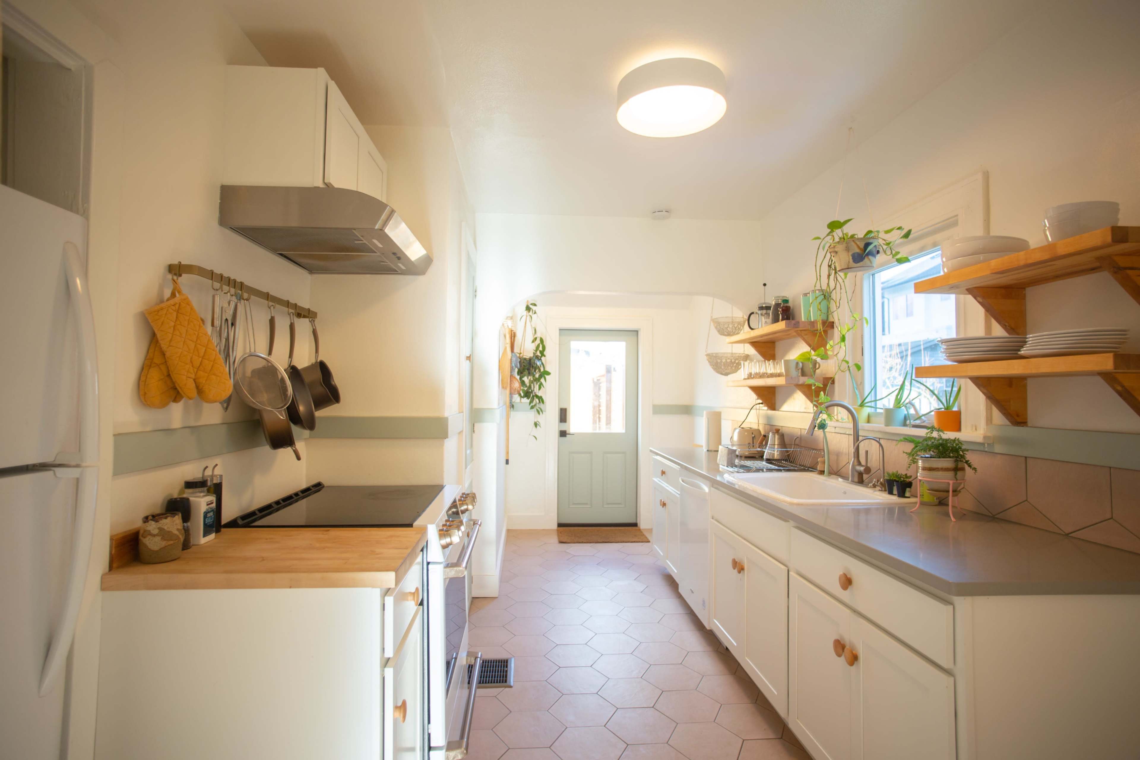 The kitchen features a white color scheme with wooden accents, a stove, and a sink under a window, along with shelves holding dishes and plants.