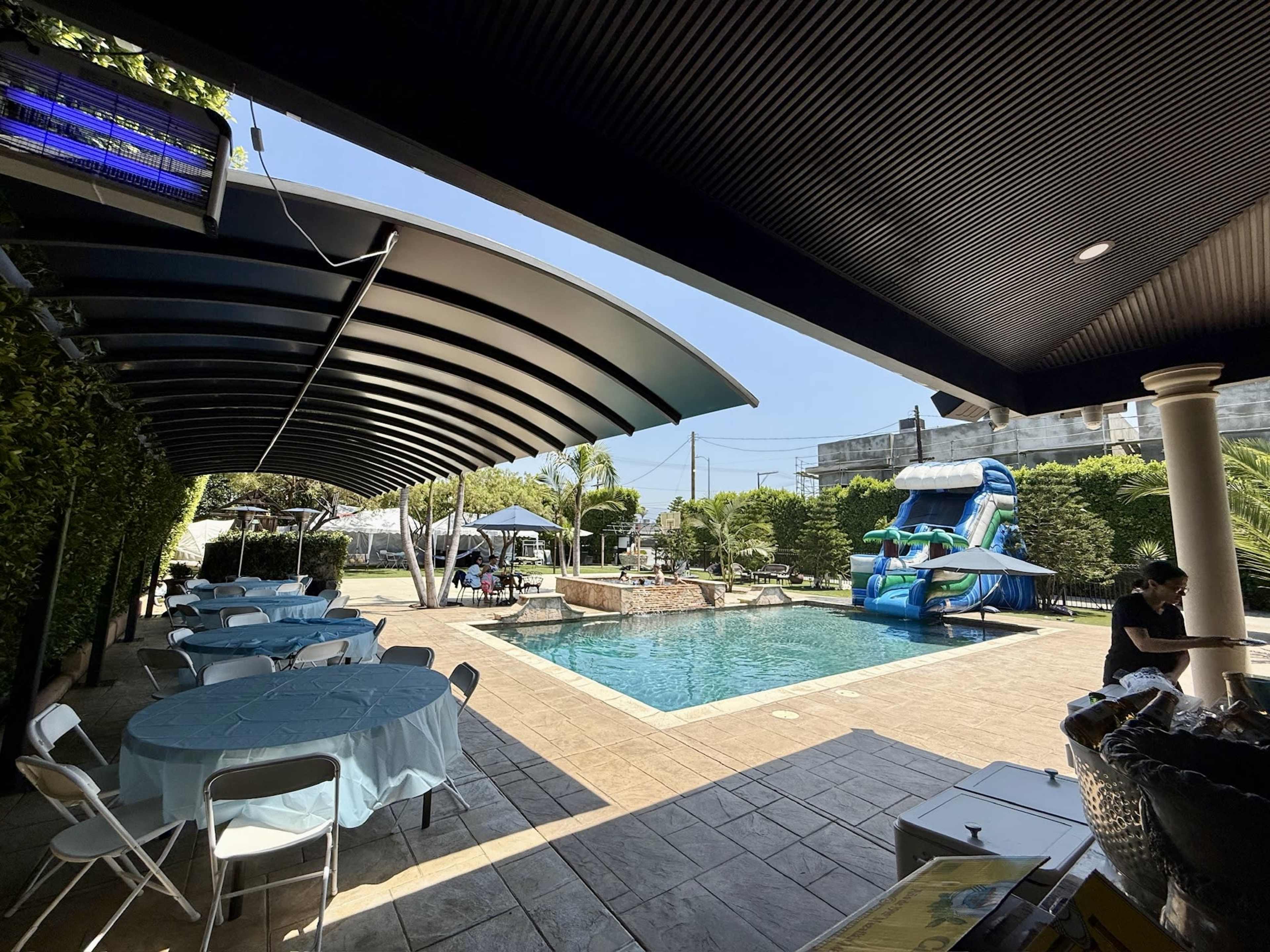 The image shows a swimming pool area with a water slide, surrounded by greenery and tables set for an event.