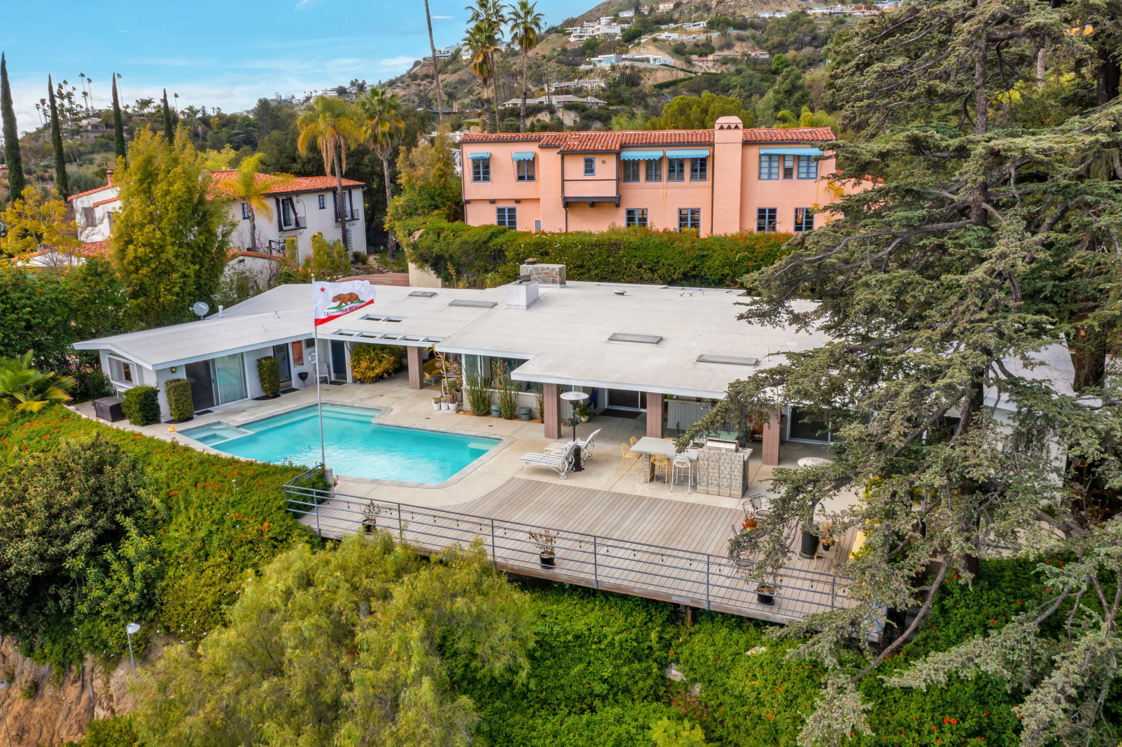 The image shows a modern home with a swimming pool, surrounded by greenery and overlooking a hillside with additional residential buildings in the background.