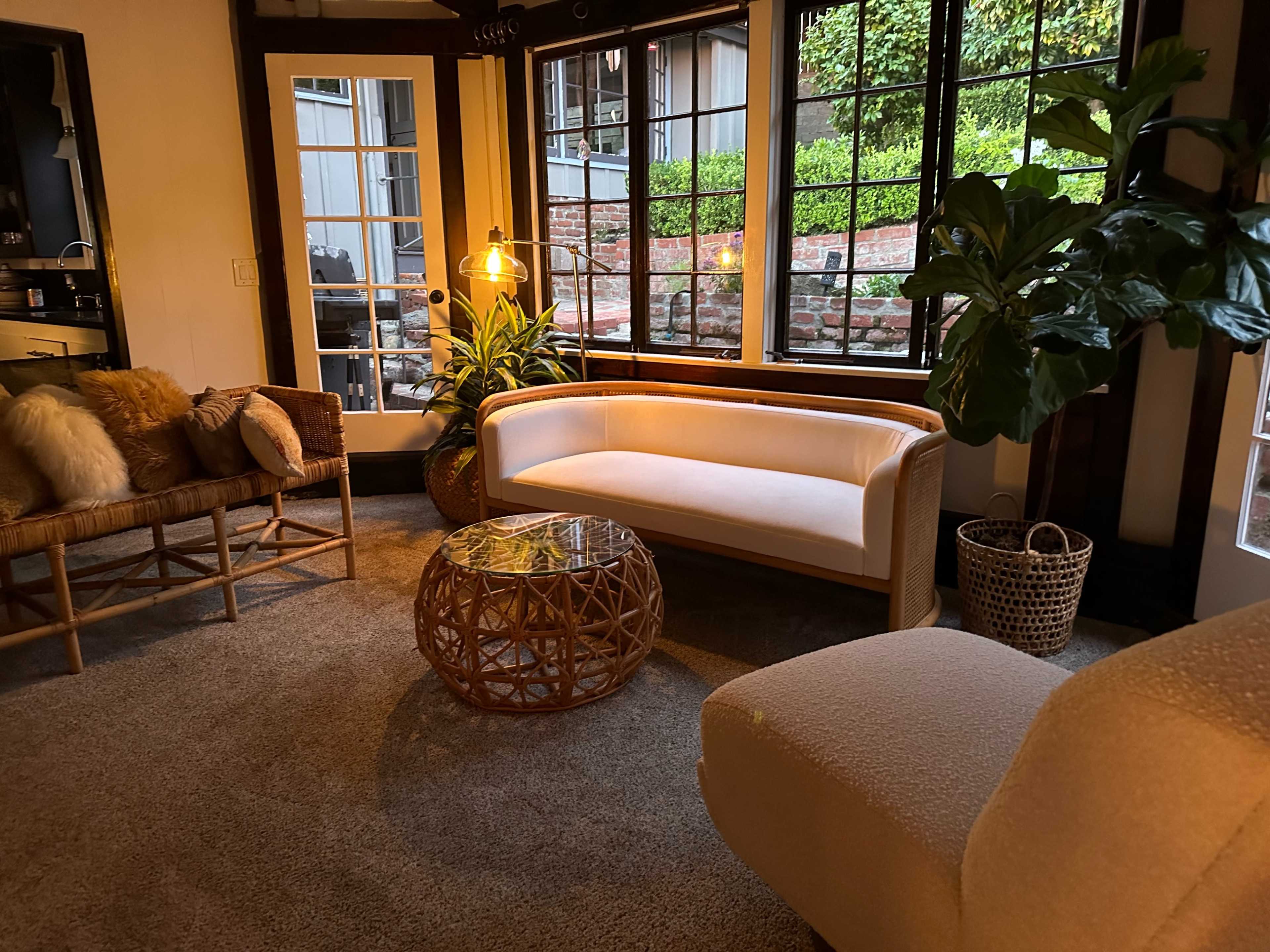 A cozy living room with a light-colored couch, a woven coffee table, and large windows allowing natural light to illuminate the space.