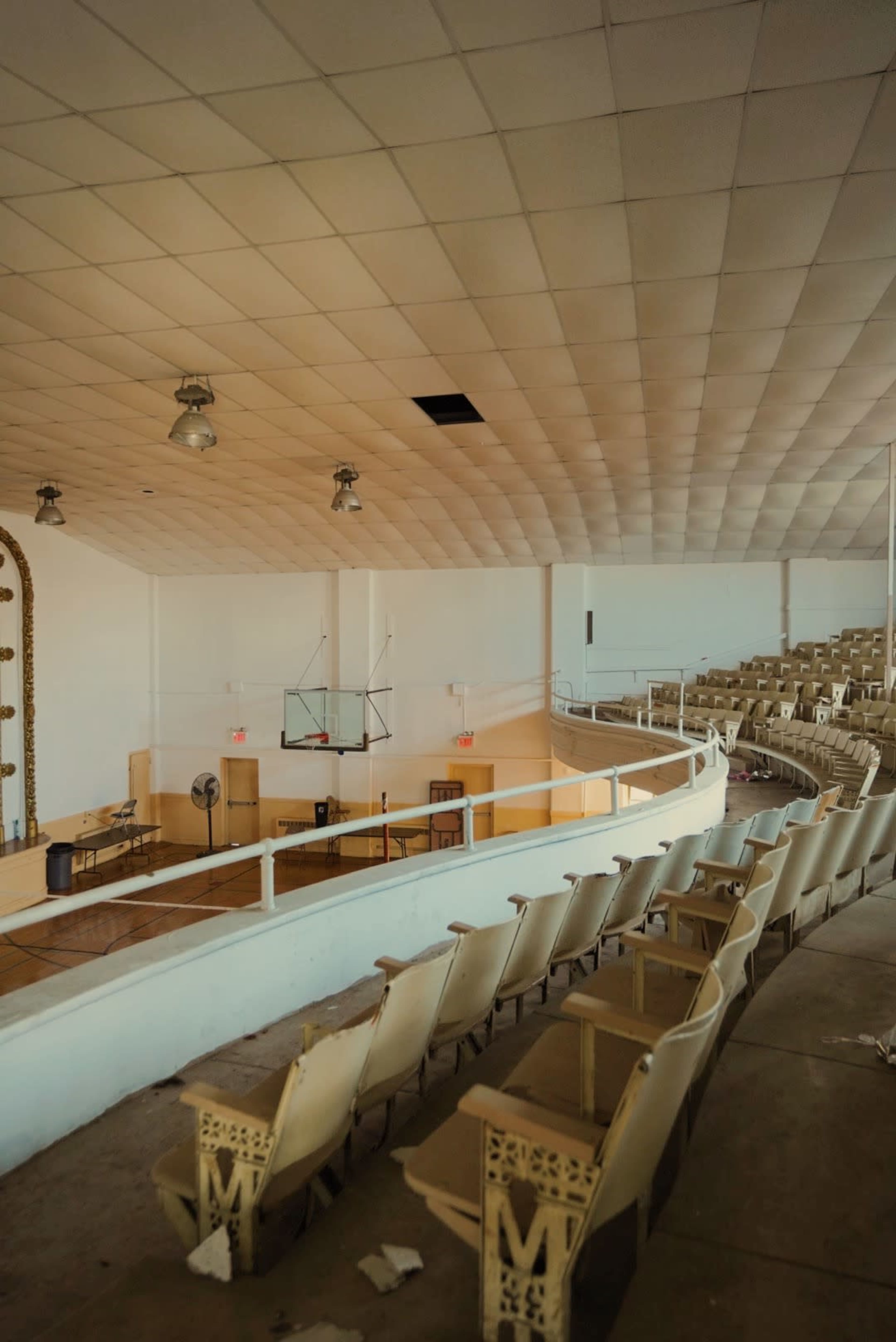 An abandoned auditorium with empty rows of seats and a basketball hoop visible in the background.
