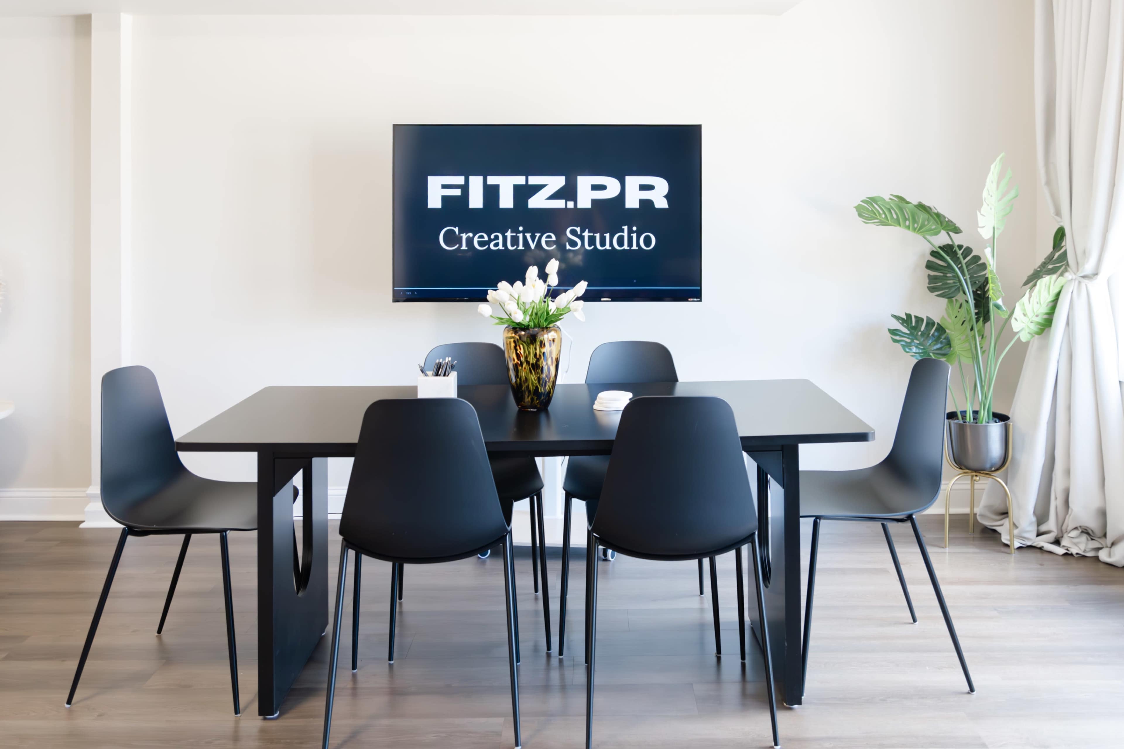A modern conference room features a black table surrounded by six chairs, with a large screen displaying "FITZ.PR Creative Studio" and a potted plant nearby.