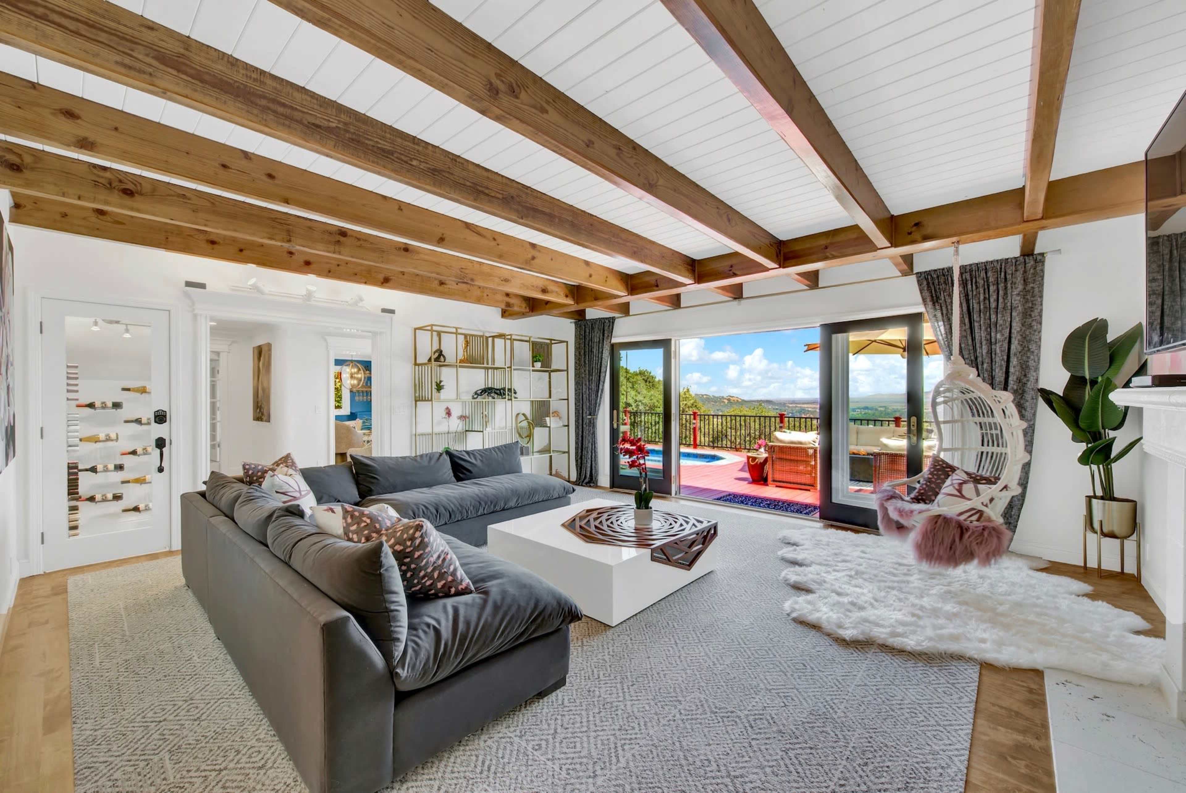 A living room with wooden beams, a large dark sofa, a white coffee table, and sliding doors that open to a balcony with outdoor seating and a view.