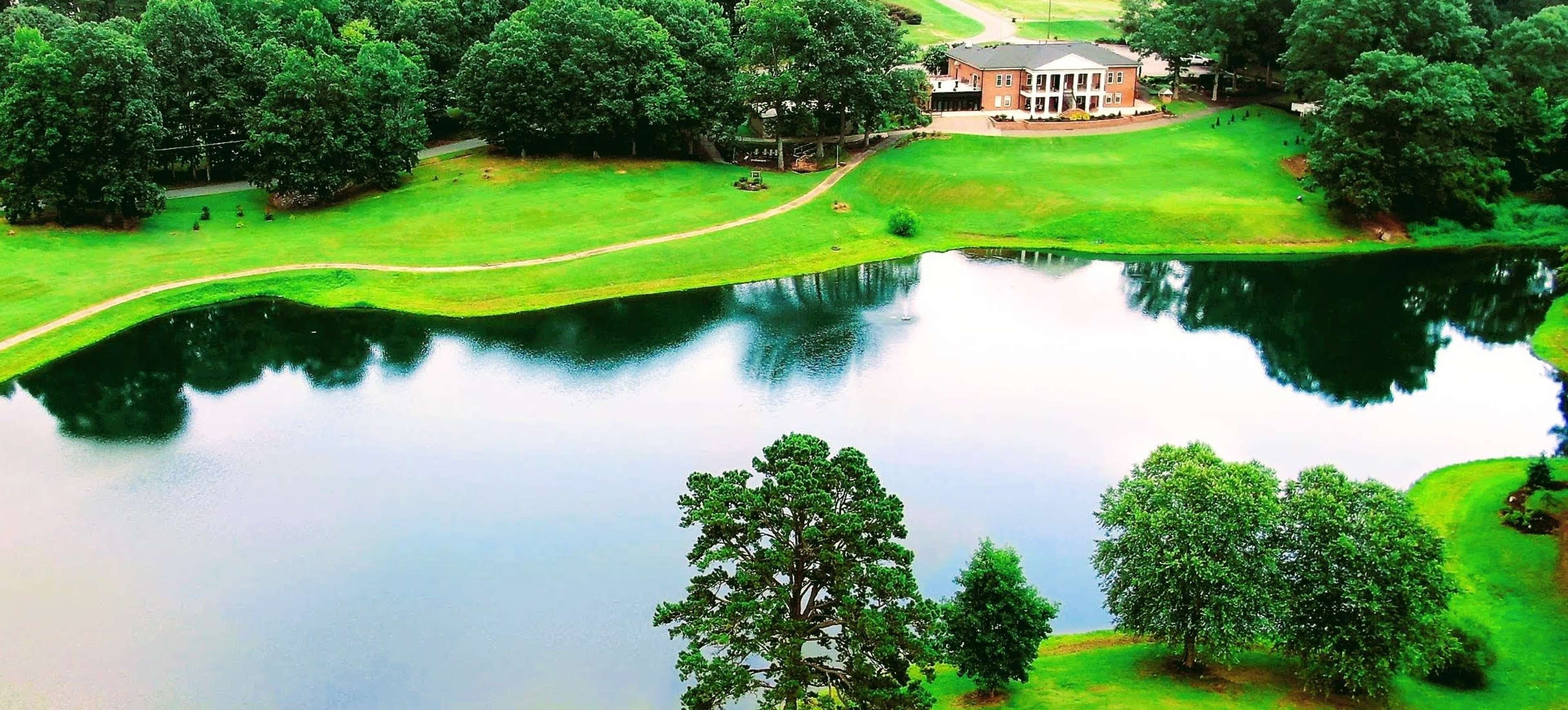 A clear lake reflects a lush green landscape, with a building and trees visible along the water's edge.