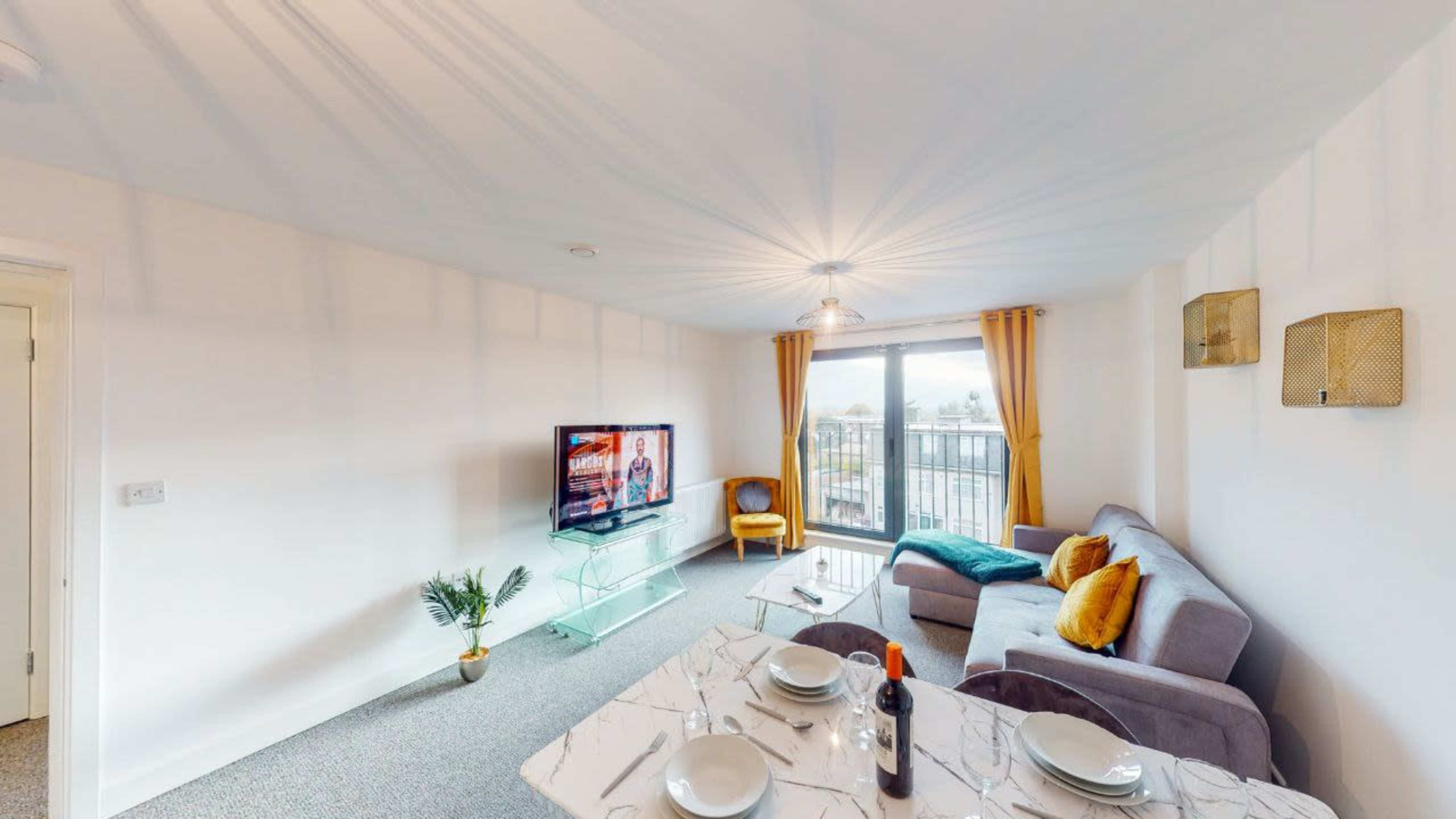 A modern living area features a television, a glass coffee table, and a sofa with accent pillows, alongside a dining area set for a meal.