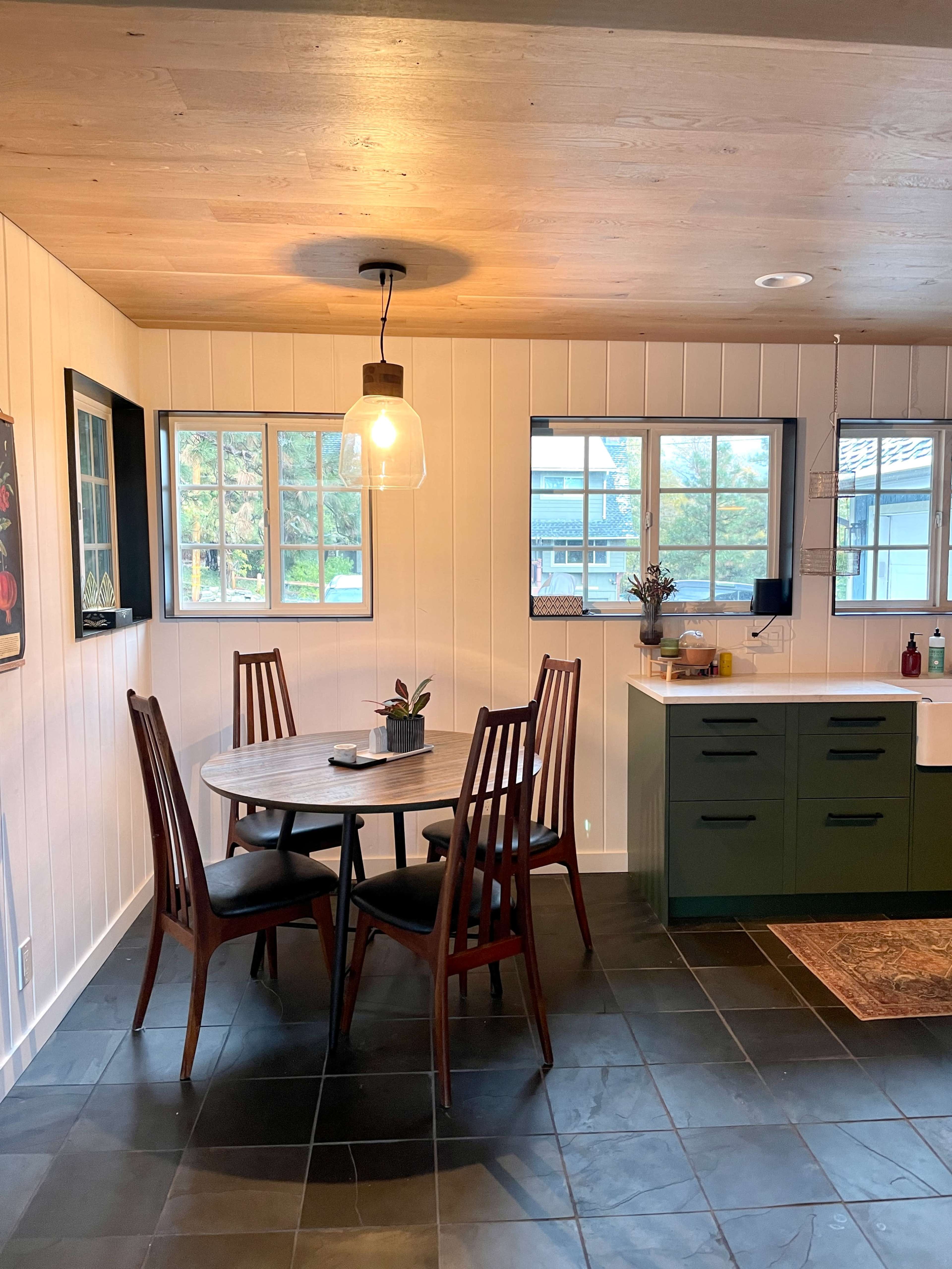 The image shows a kitchen with a round dining table surrounded by four chairs, large windows providing natural light, and a green cabinetry setup.