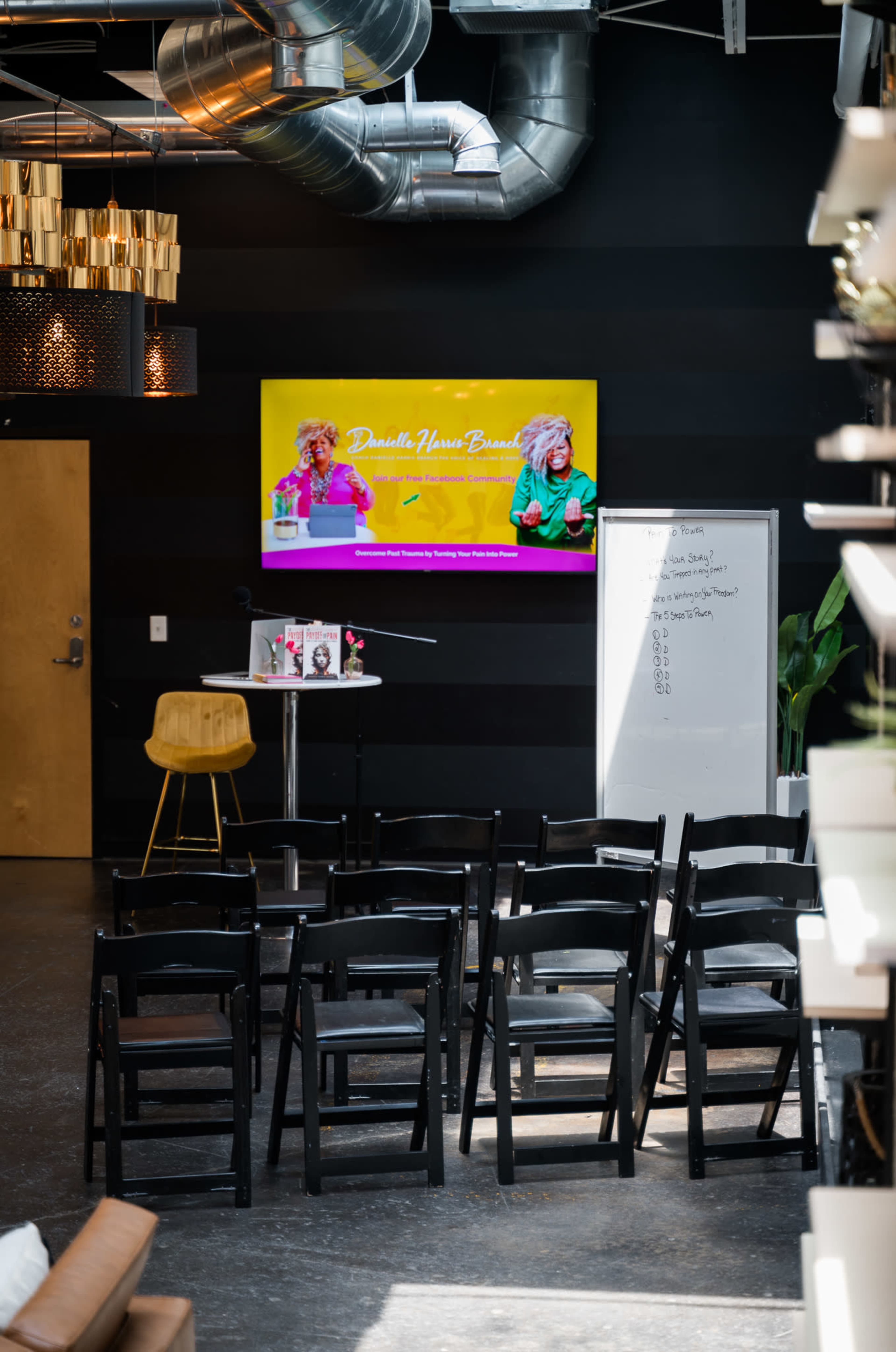 A modern workspace features rows of black chairs facing a large screen displaying a colorful presentation, with a whiteboard and a small table nearby.