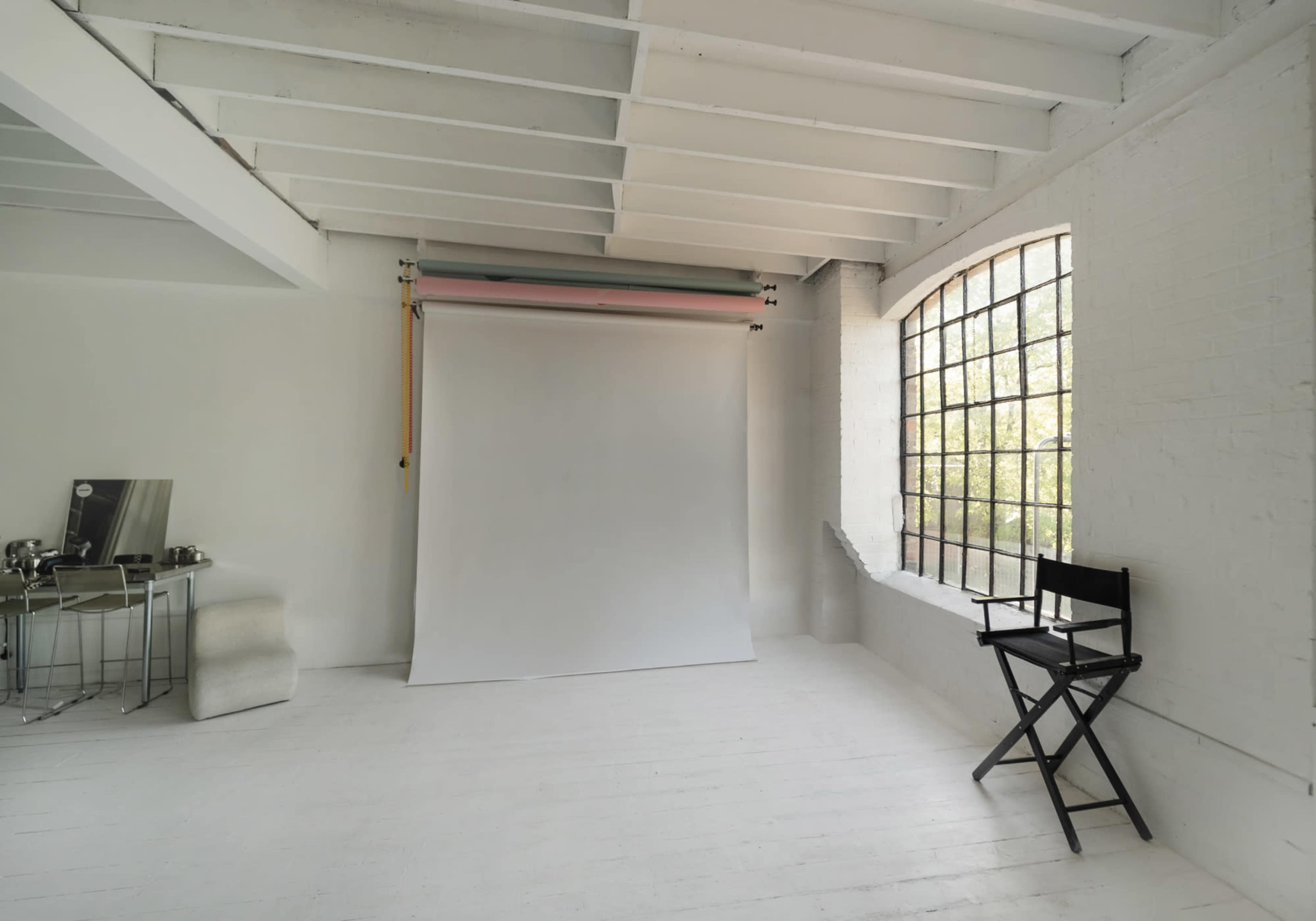 The image shows a minimalist studio space with a large window, white walls, and a backdrop setup beside a director's chair.