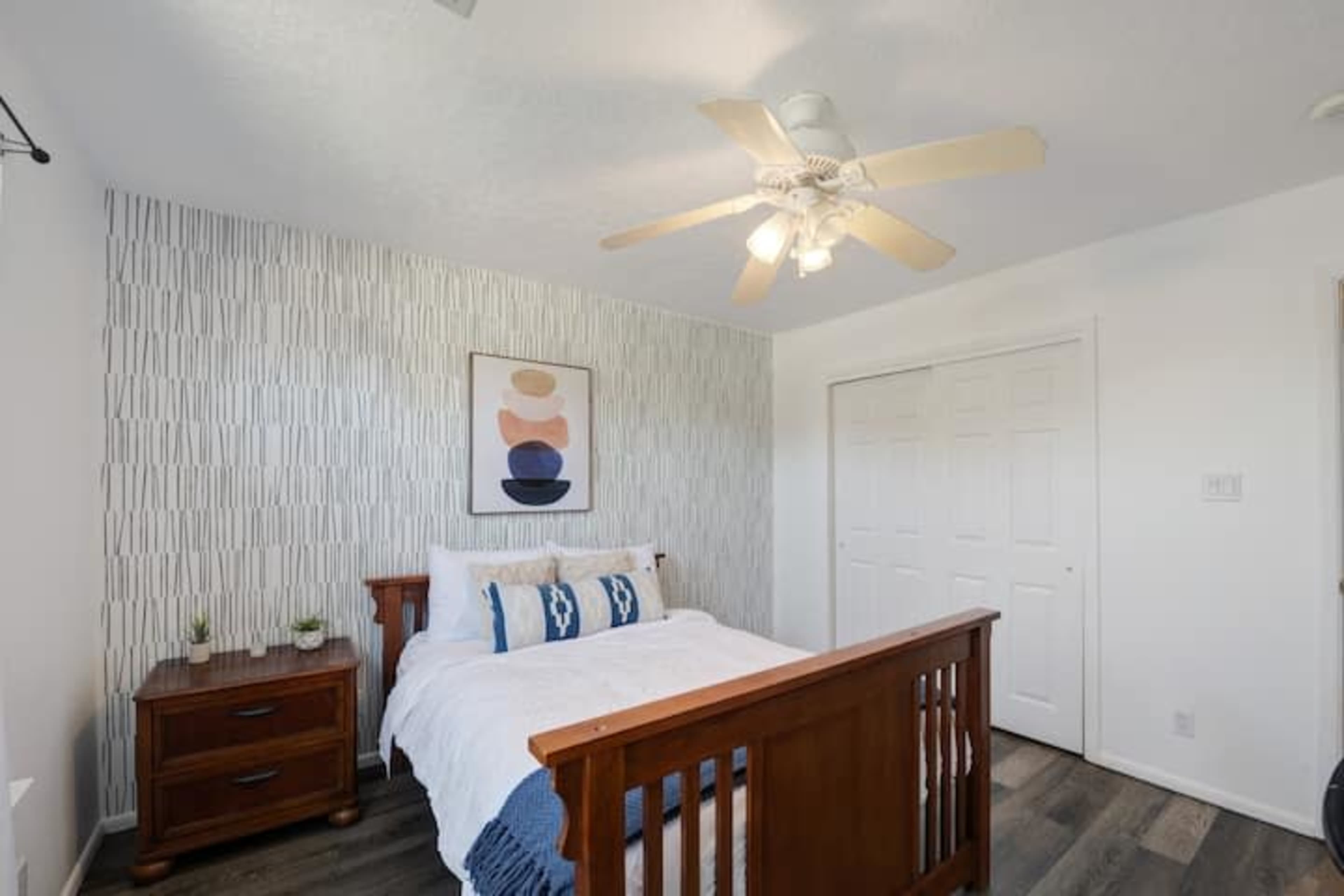A bedroom featuring a wooden bed frame, a light-colored wall with a patterned design, and a ceiling fan with bright lights.