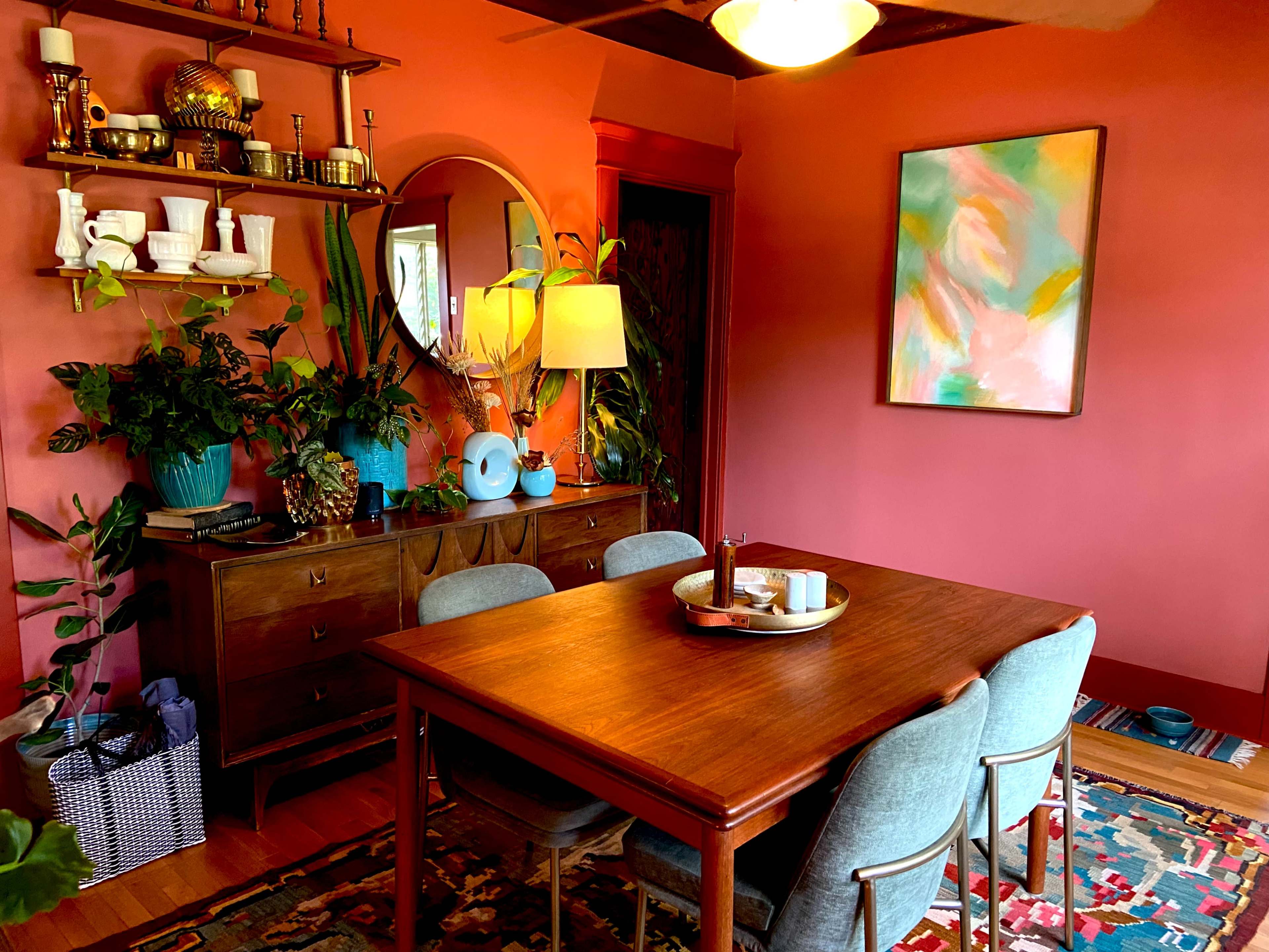 The image depicts a dining area with a wooden table surrounded by four chairs, a sideboard adorned with plants, and a colorful painting on the wall.