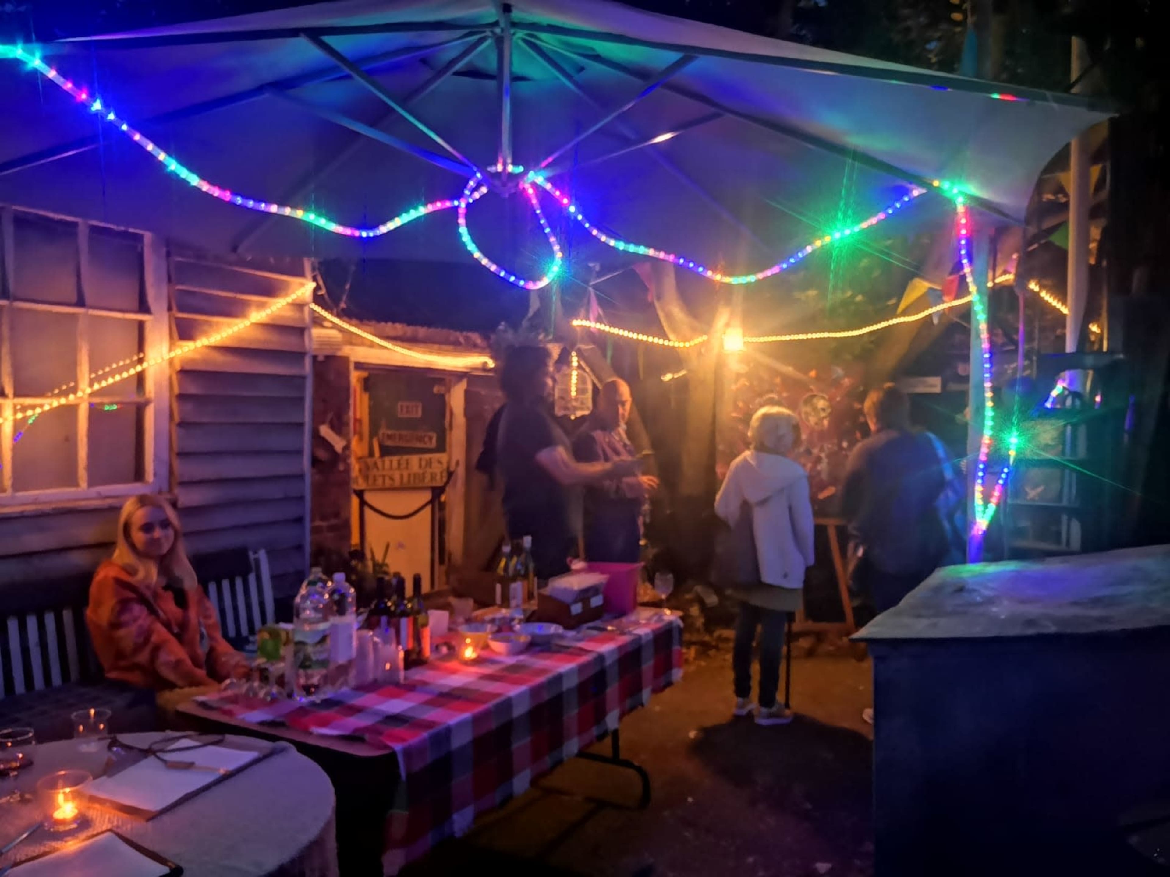 A group of people gathers under a lit tent adorned with colorful lights in a backyard setting at night.