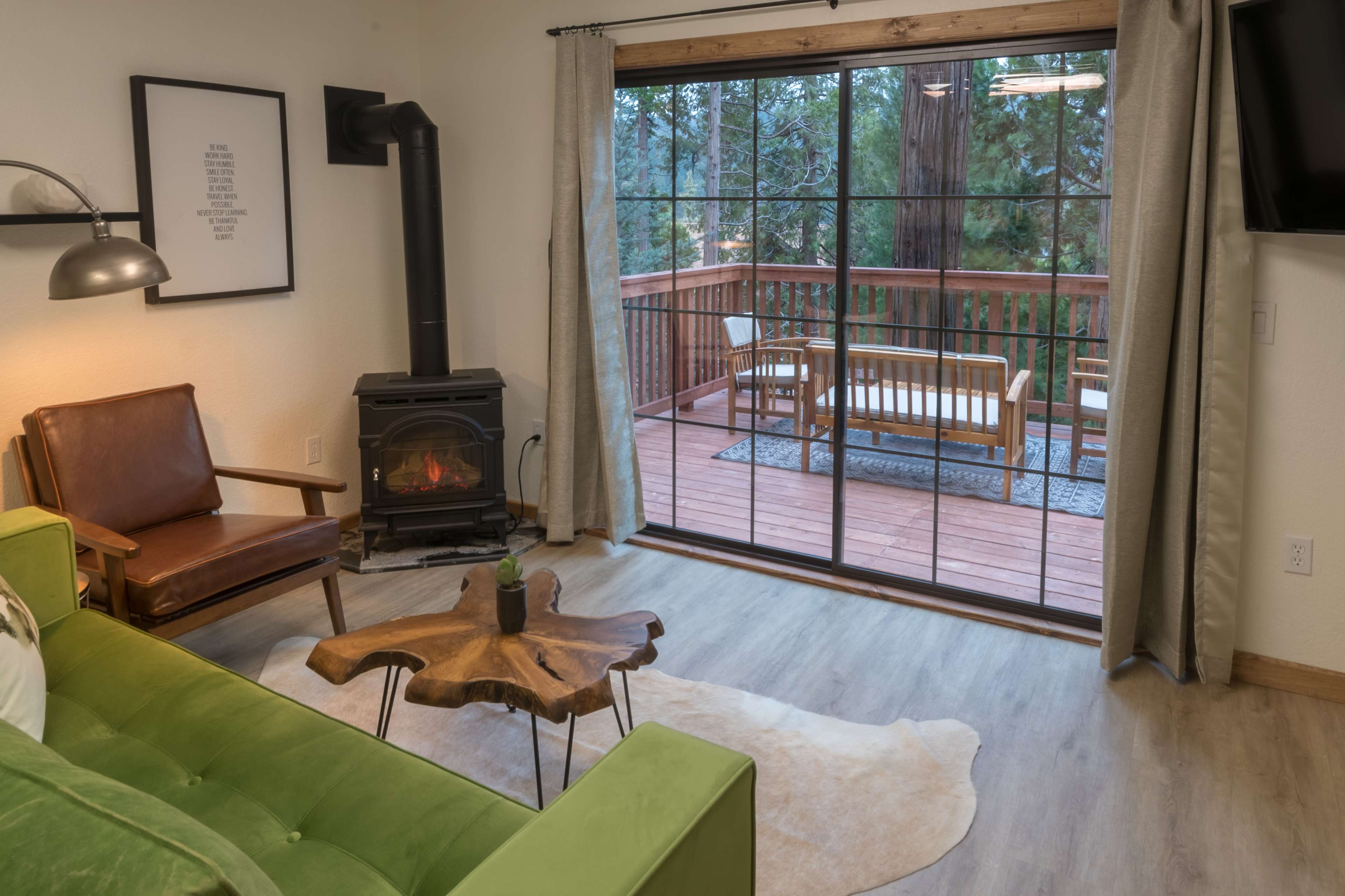 A cozy living room features a green sofa, a wooden table, and a wood stove, with a large window showing a deck and trees outside.