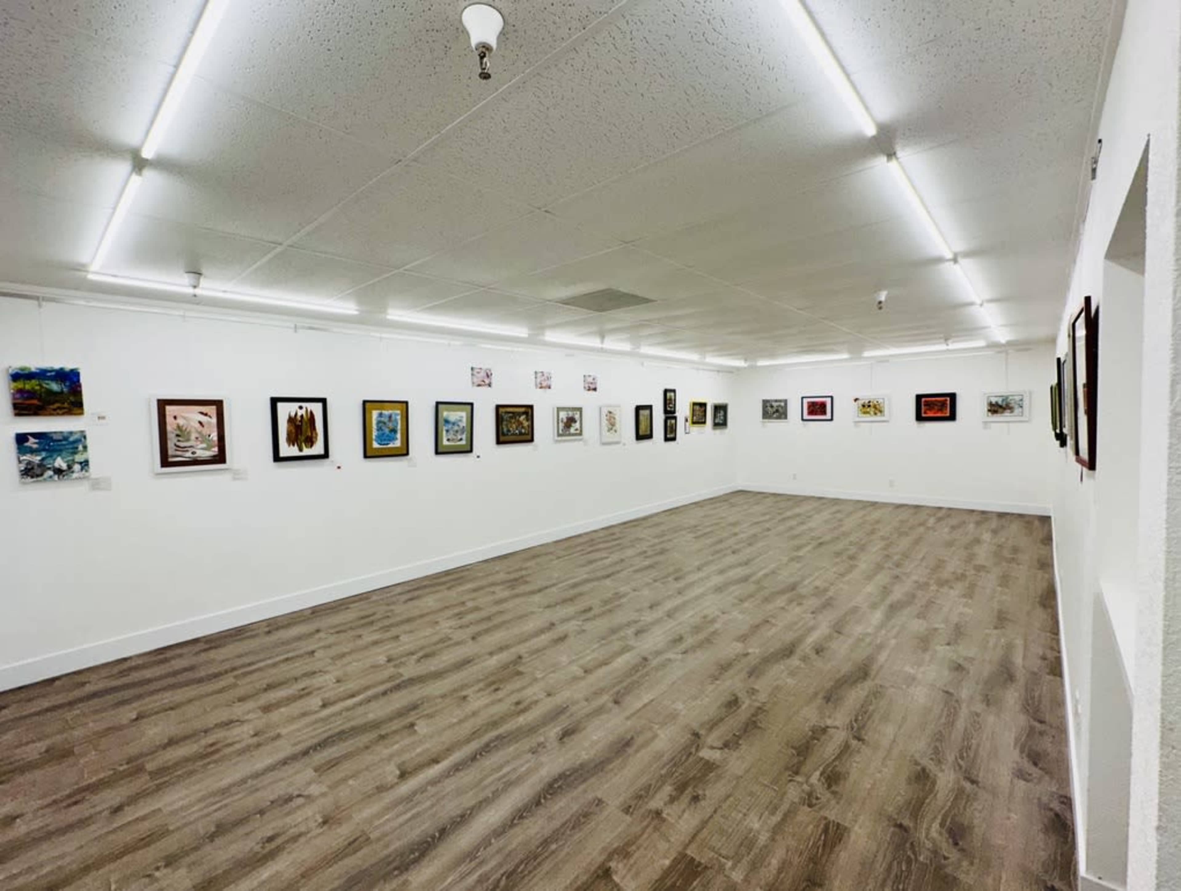 A white-walled art gallery with wooden flooring, featuring a series of framed artworks hanging evenly spaced along the walls.