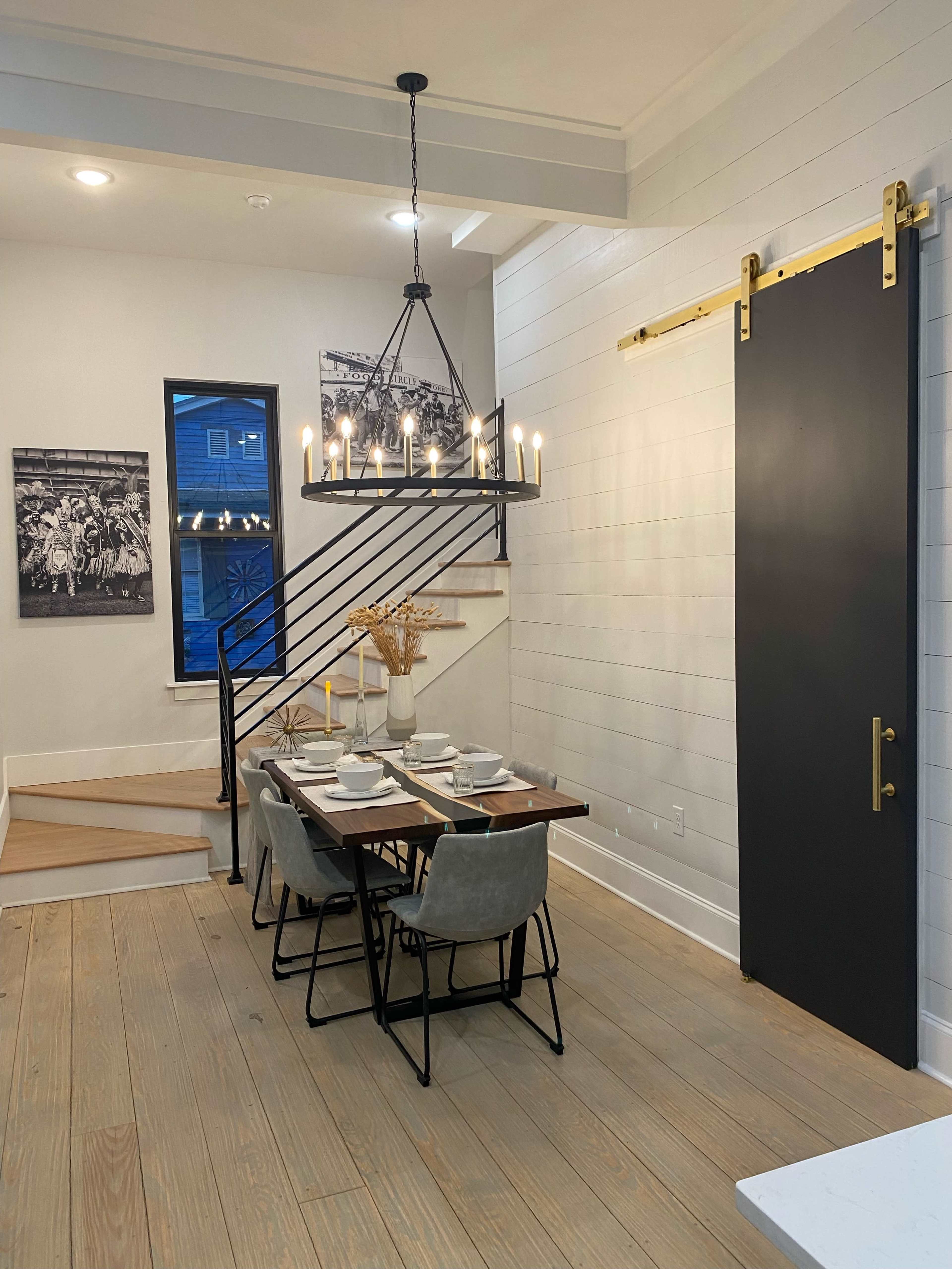 A modern dining area features a wooden table set for dinner under a chandelier, with a sliding door and staircase in the background.