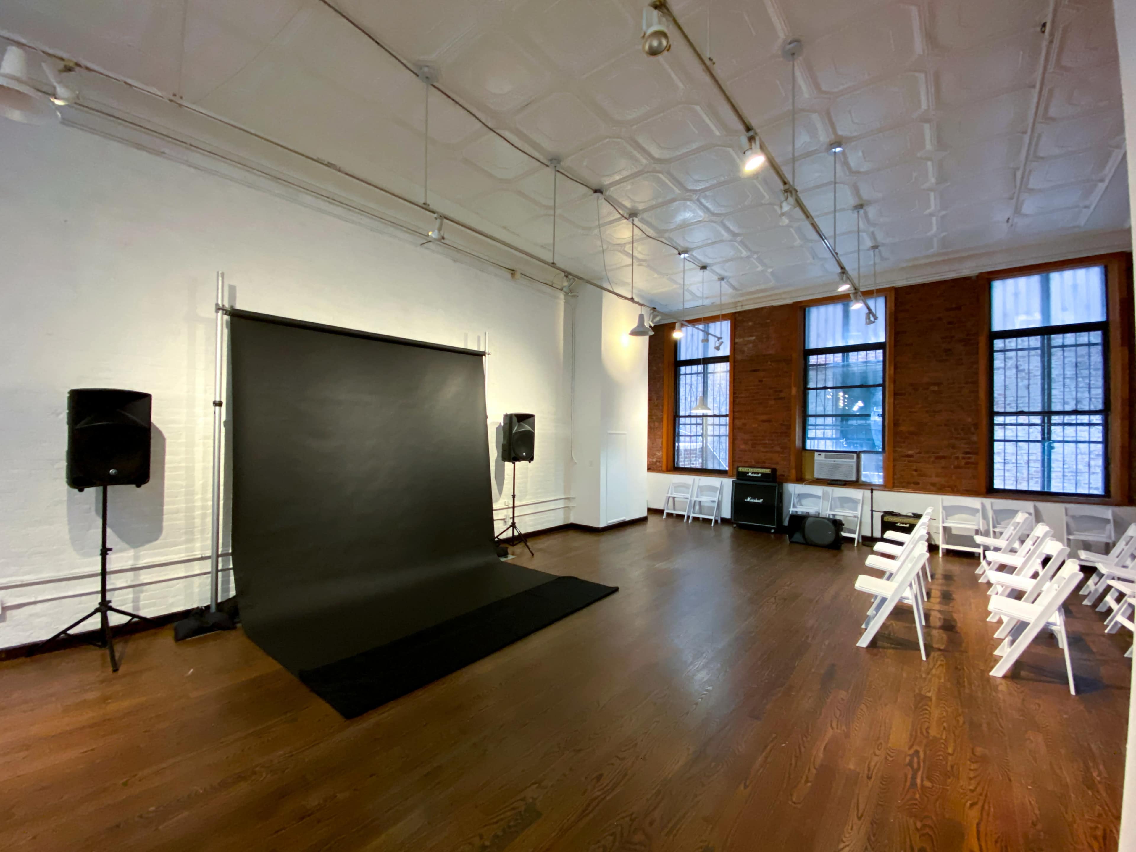 The image shows a spacious room with a black backdrop setup, flanked by speakers and arranged chairs facing the backdrop.