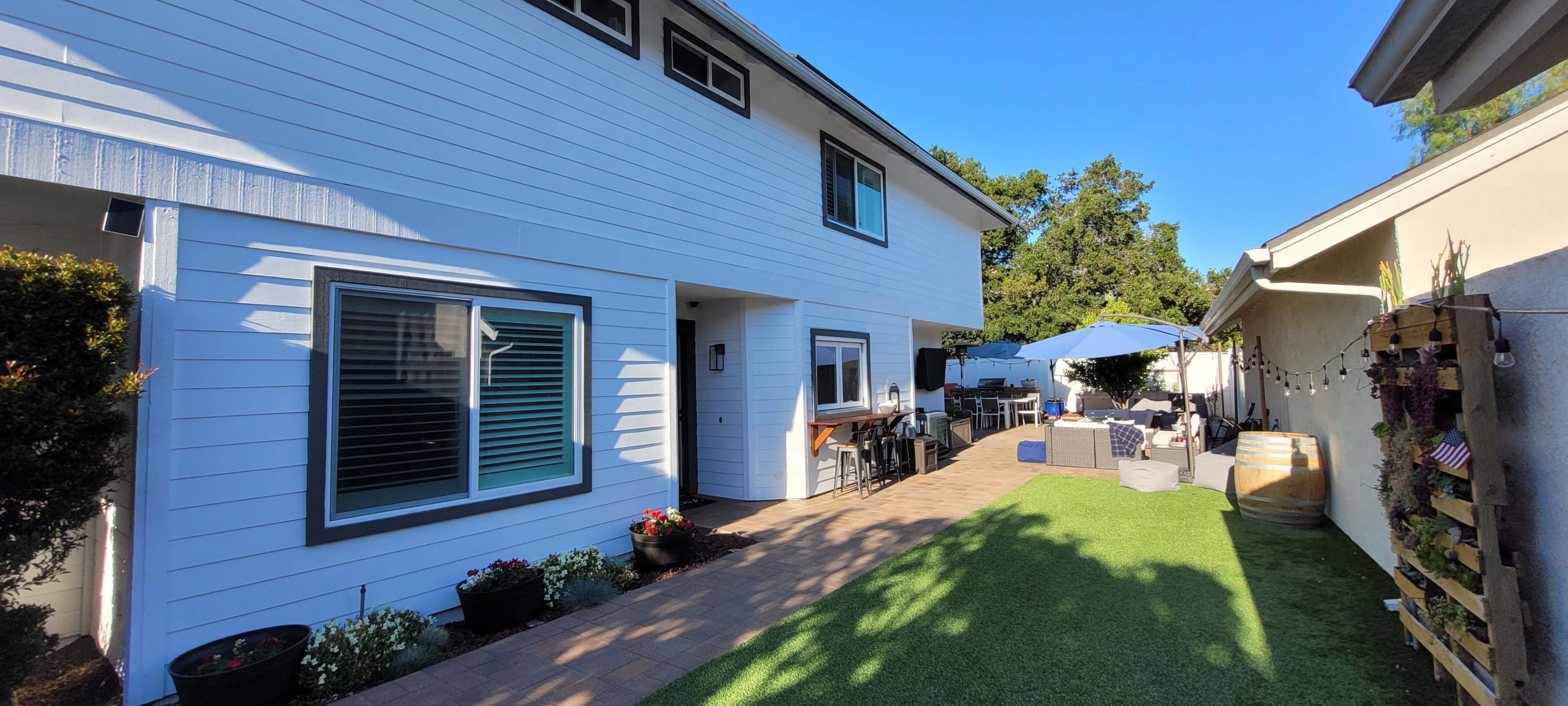 The image shows a backyard area with a lawn, a patio set under an umbrella, and a house with a white exterior and large windows.