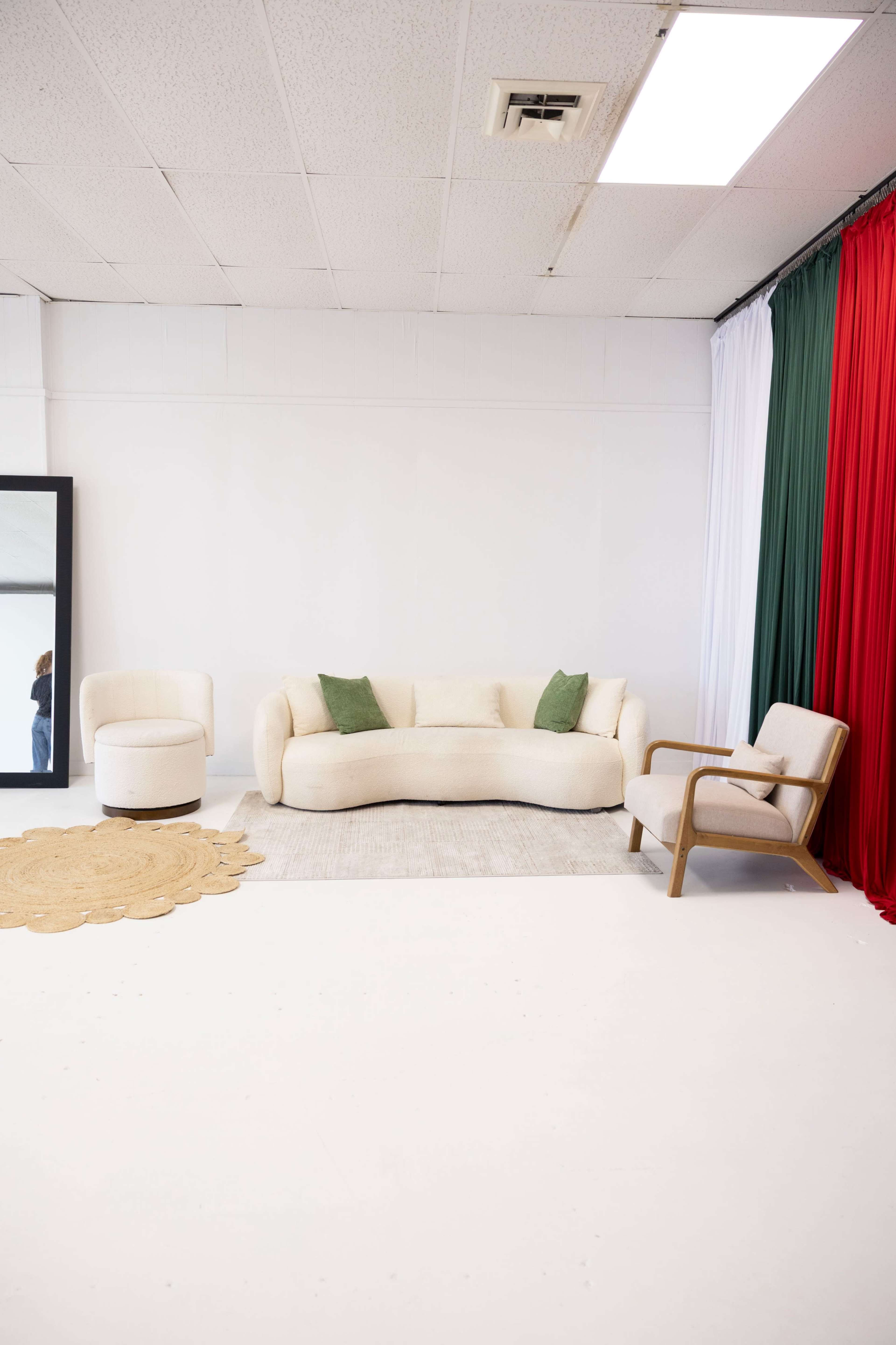 The image depicts a minimalist interior space featuring a neutral-toned sofa with green pillows, a matching armchair, a round ottoman, and a jute rug, alongside a large mirror and colorful drapes of red, white, and green.