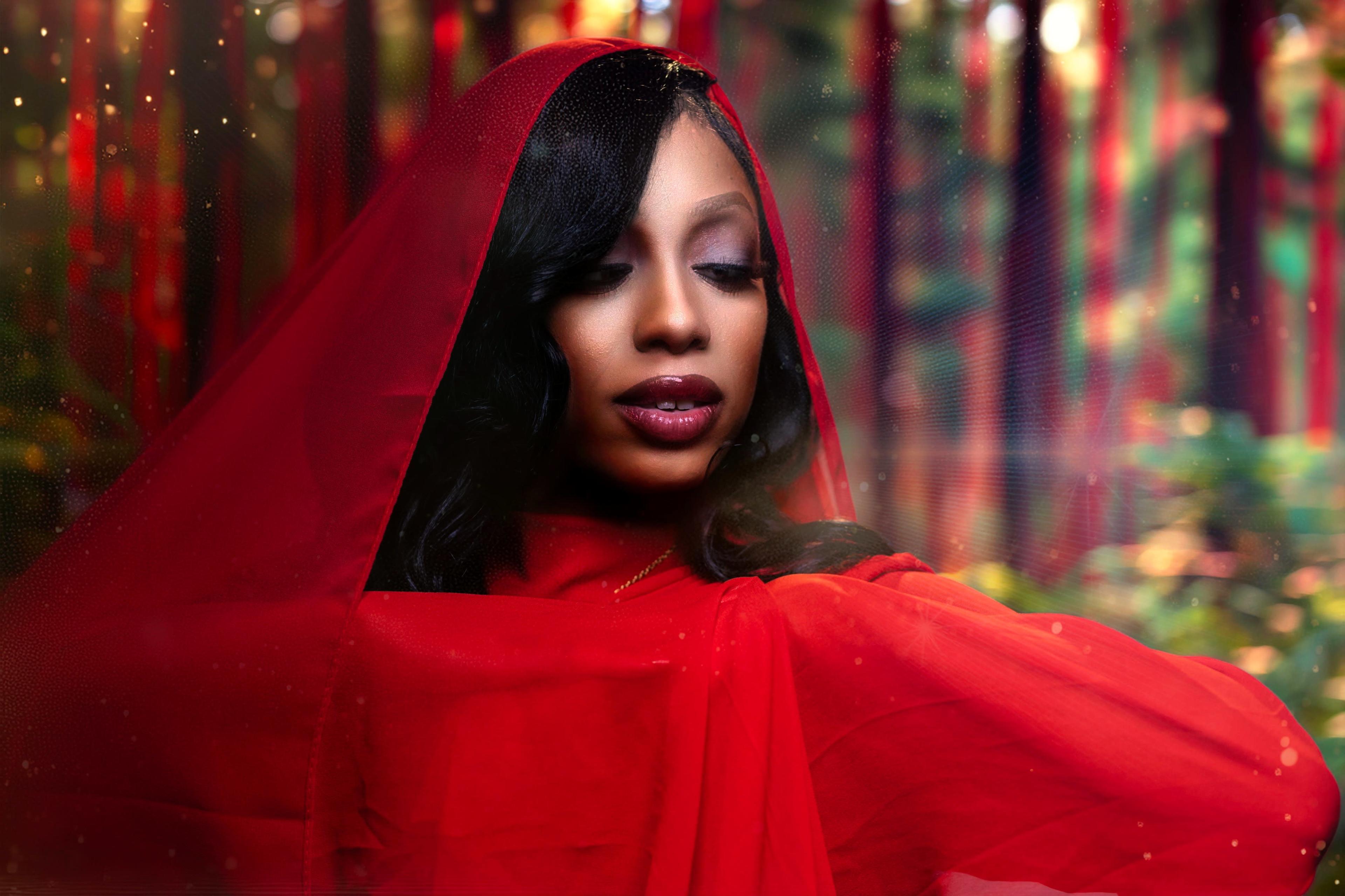 A woman wearing a flowing red veil poses against a blurred, colorful forest background.