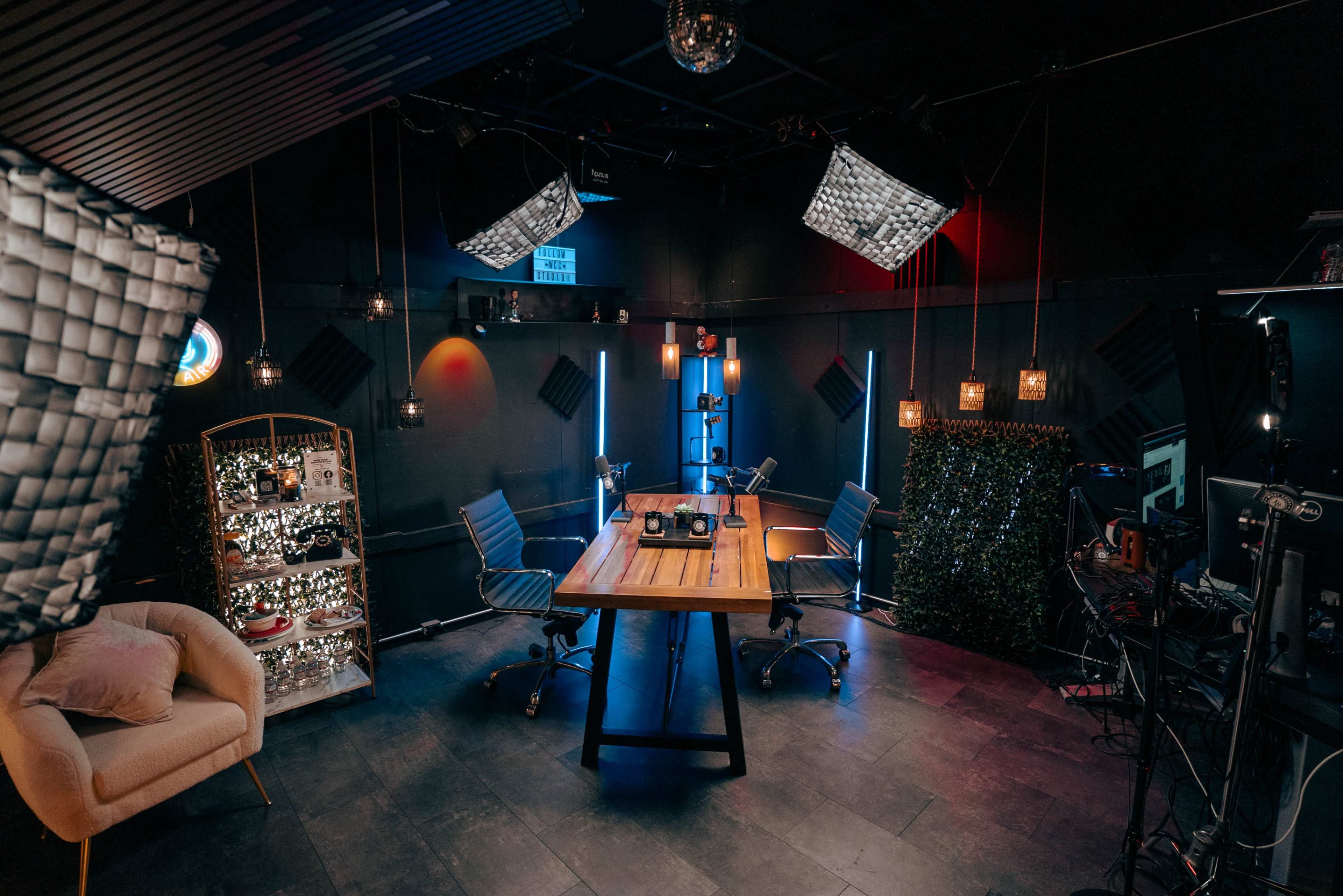 The image shows a modern recording studio with two chairs, a wooden table, and various lighting setups.