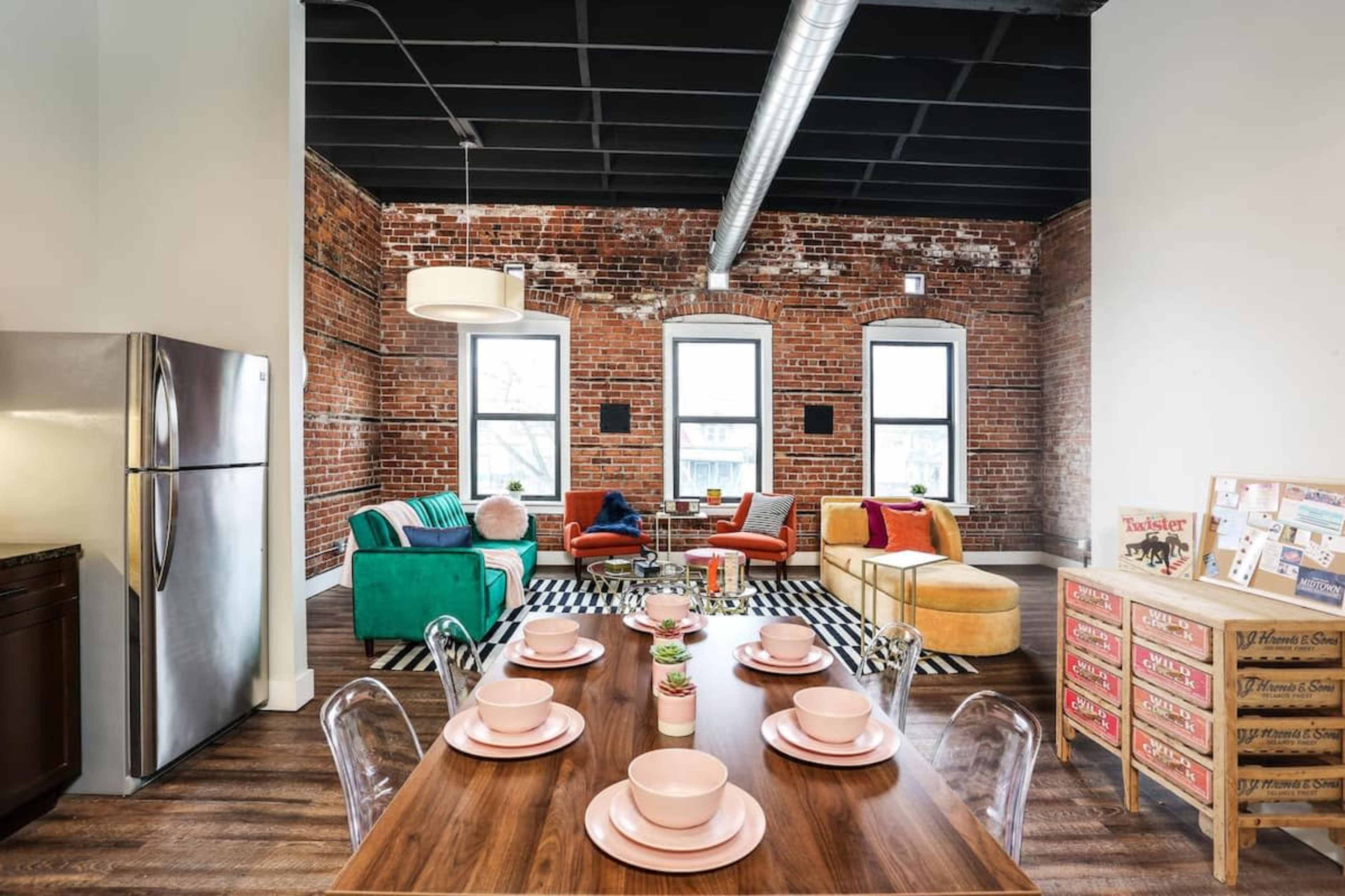 A modern living space featuring exposed brick walls, large windows, a mix of colorful furniture, and a dining table set with pink plates.