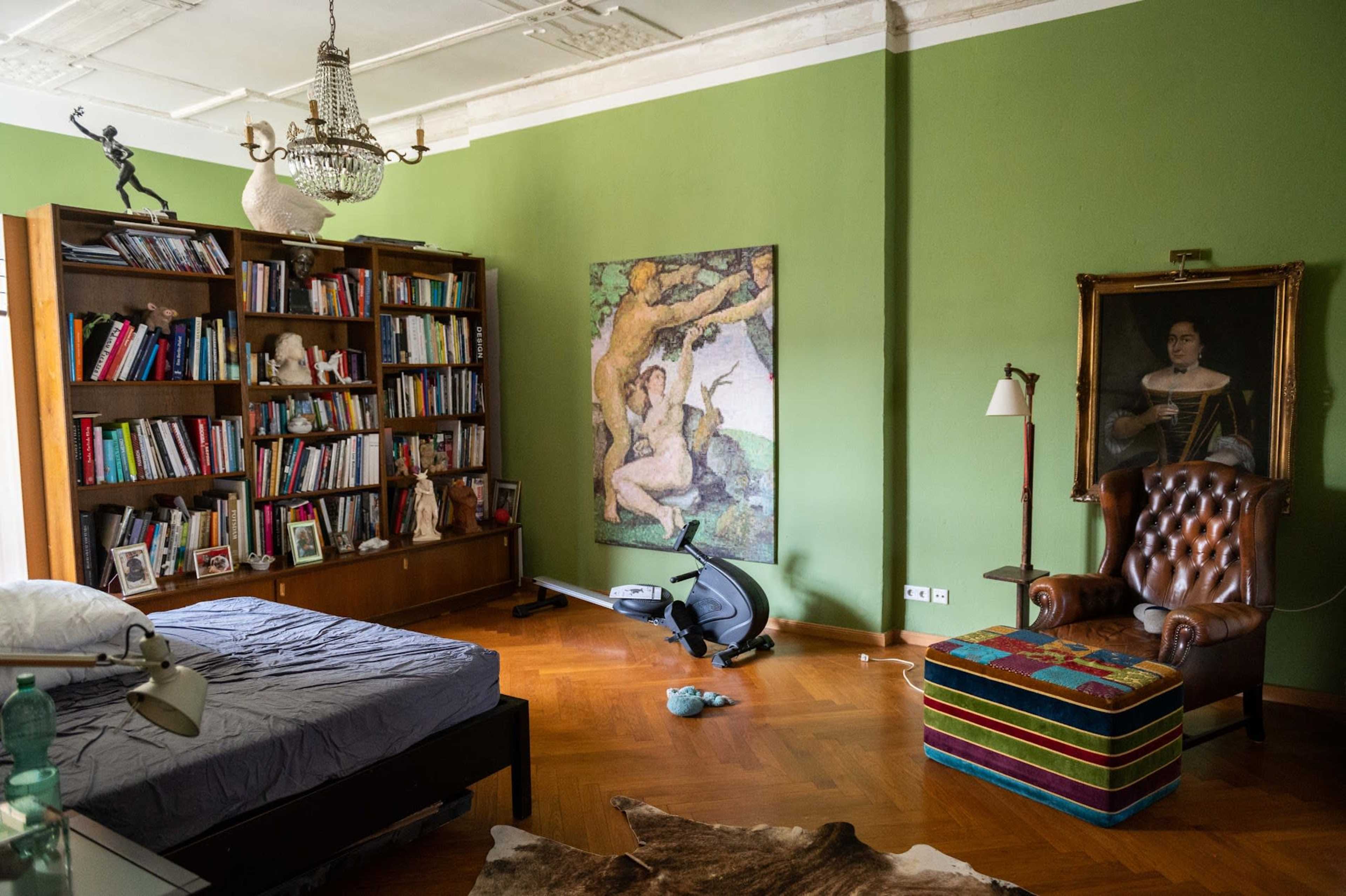 A room features a green wall, a bookshelf filled with books, a brown leather armchair, a bed with unmade sheets, and a large artwork depicting a classical scene.