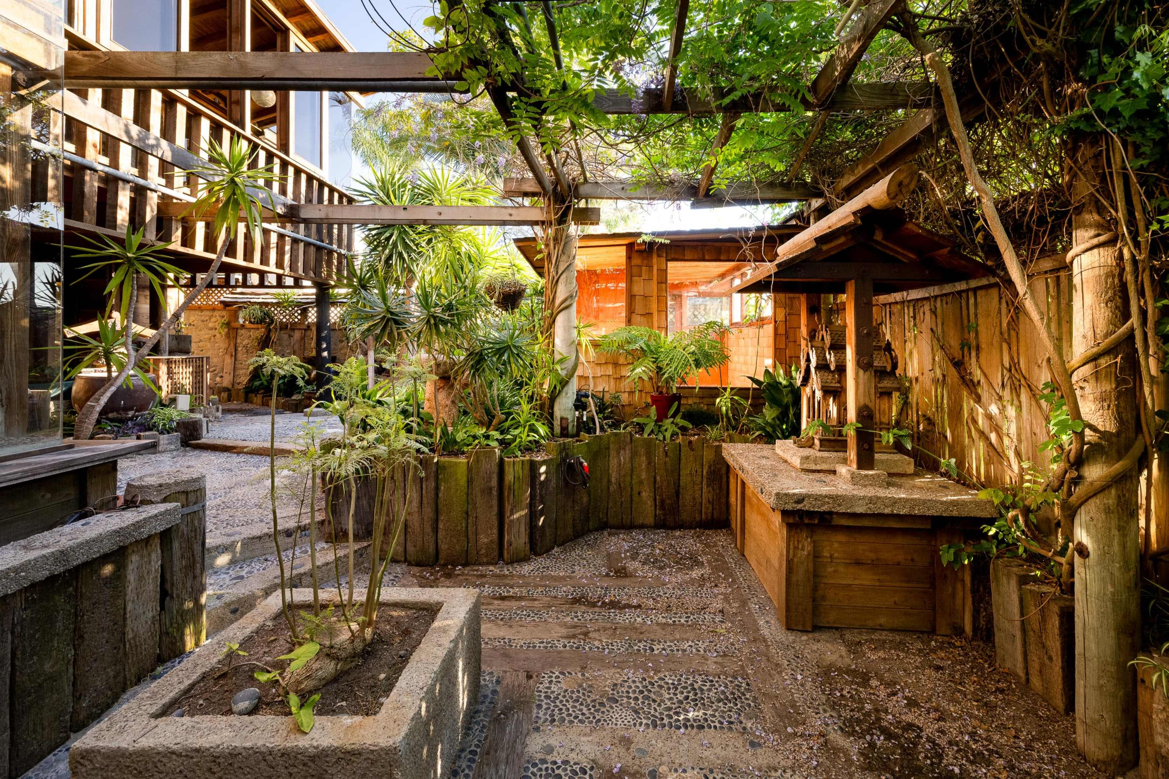 The image depicts a landscaped outdoor area with wooden structures, lush greenery, and a stone pathway leading to a decorative stone feature.