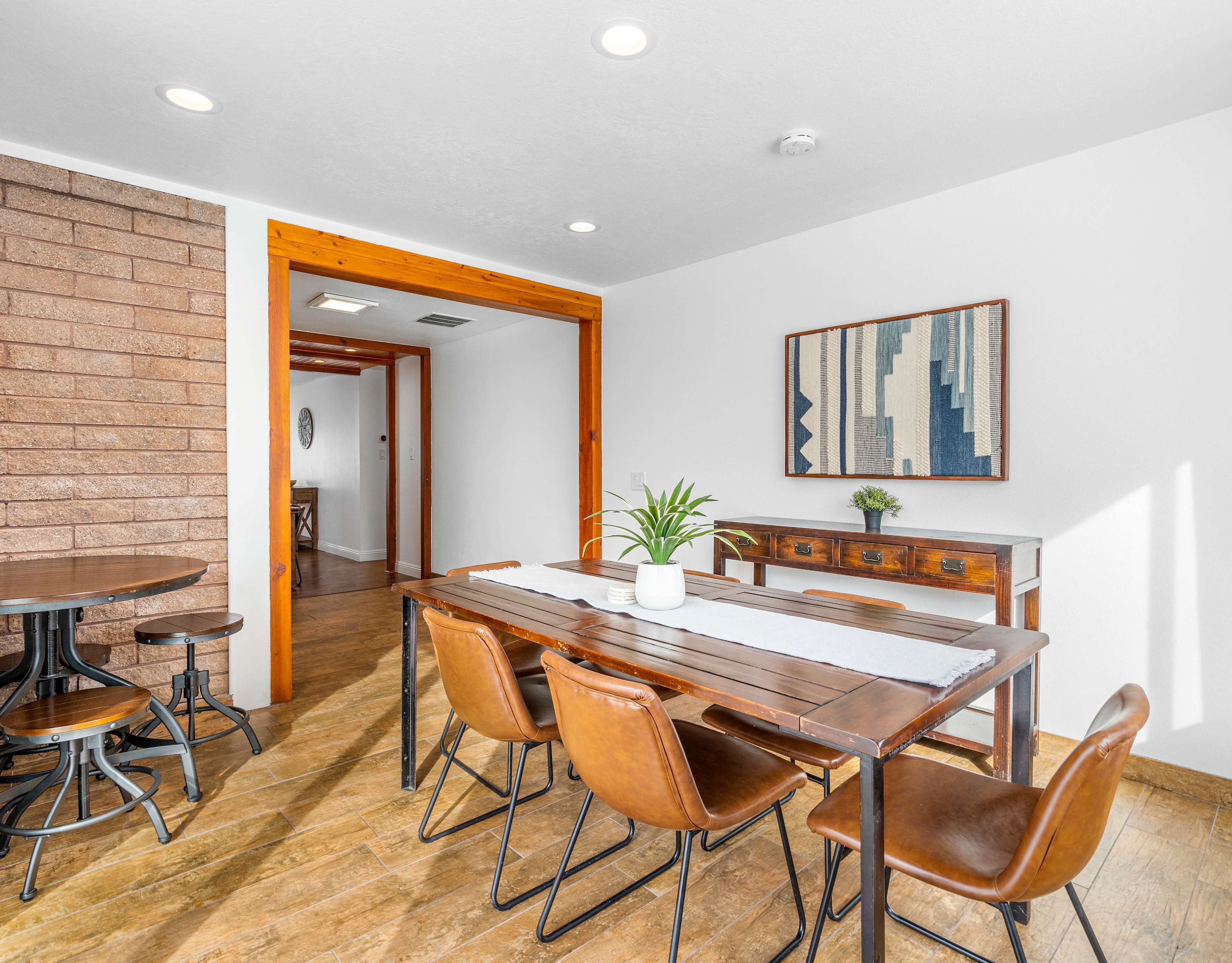 A dining area with a wooden table surrounded by leather chairs, a sideboard, and a geometric wall art, illuminated by ceiling lights.