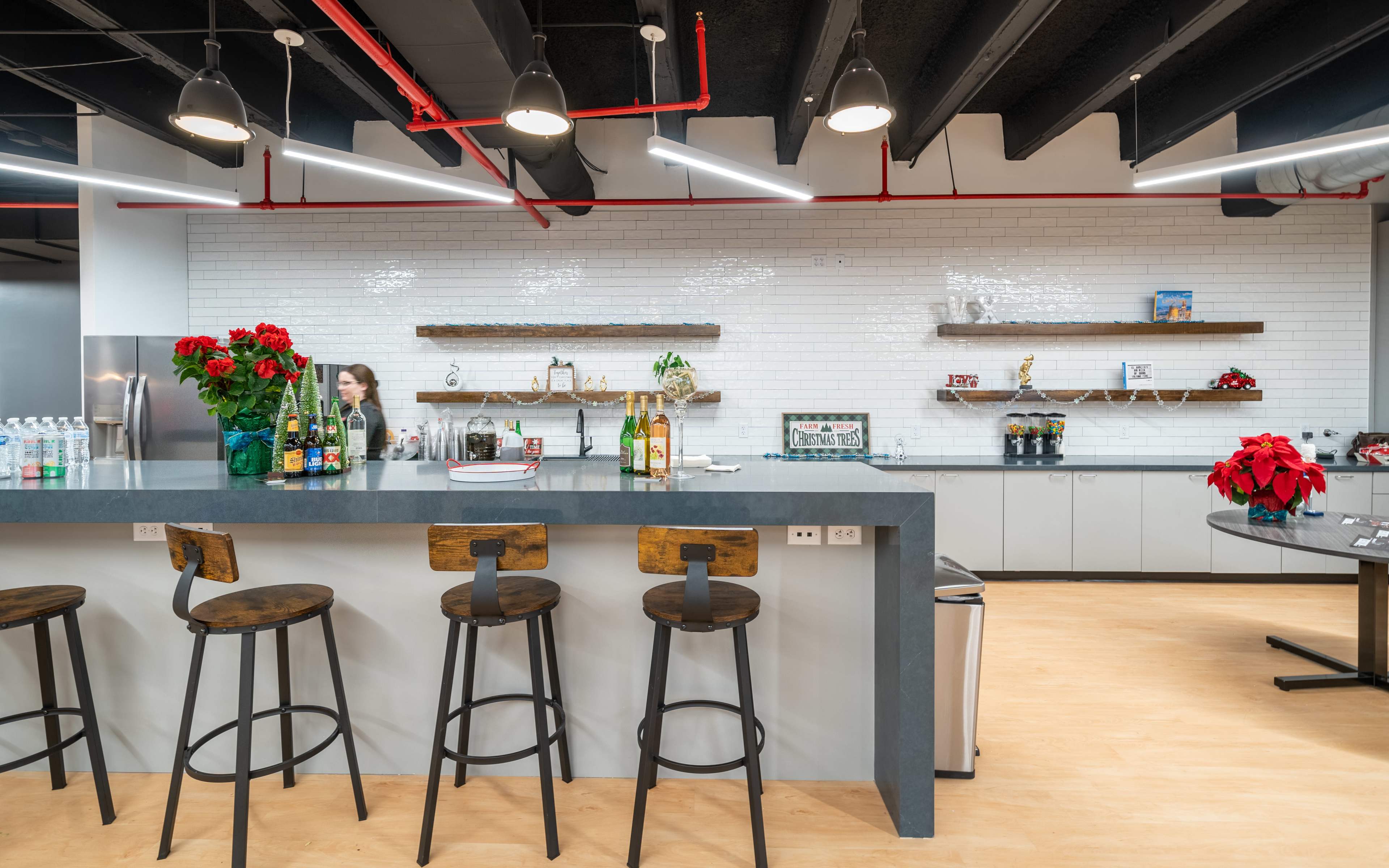 A modern office kitchen features a gray countertop with wooden barstools, a variety of drinks on display, and decorative shelves against a white tiled wall.