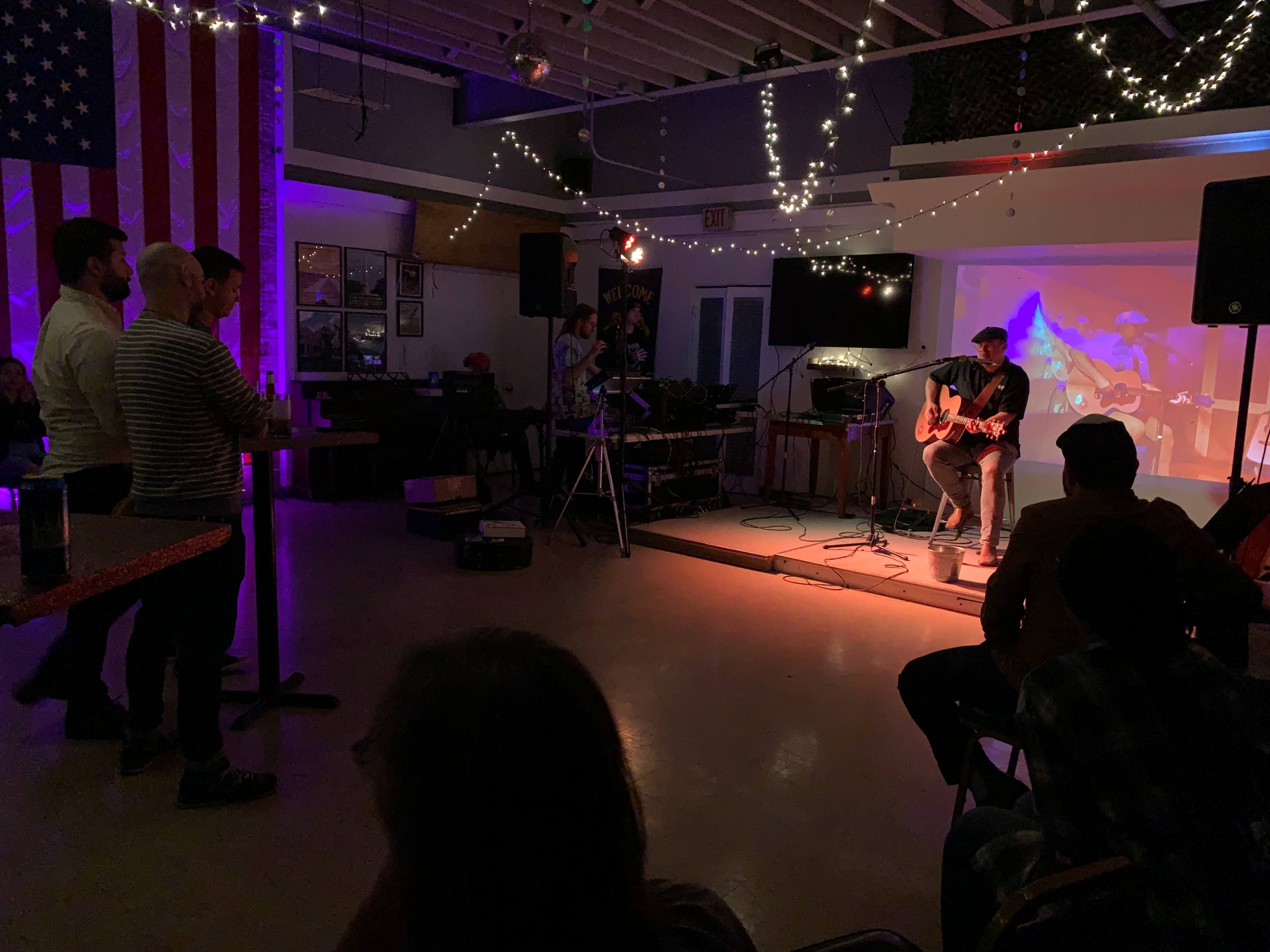 A musician plays guitar on stage while a small audience observes in a dimly lit venue adorned with string lights.