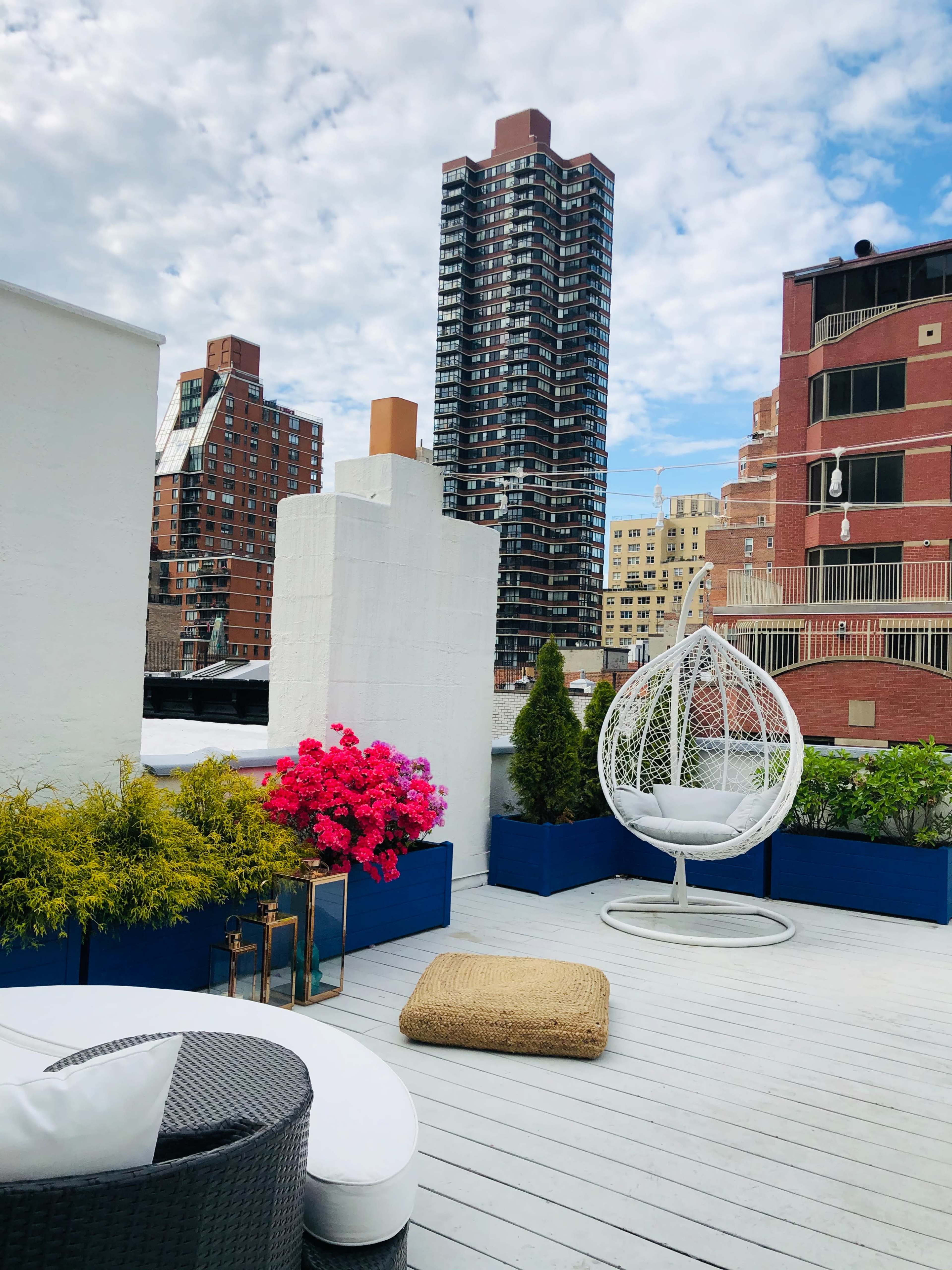 A rooftop deck featuring a white hanging chair, vibrant pink flowers, green plants, and a view of surrounding apartment buildings under a partly cloudy sky.