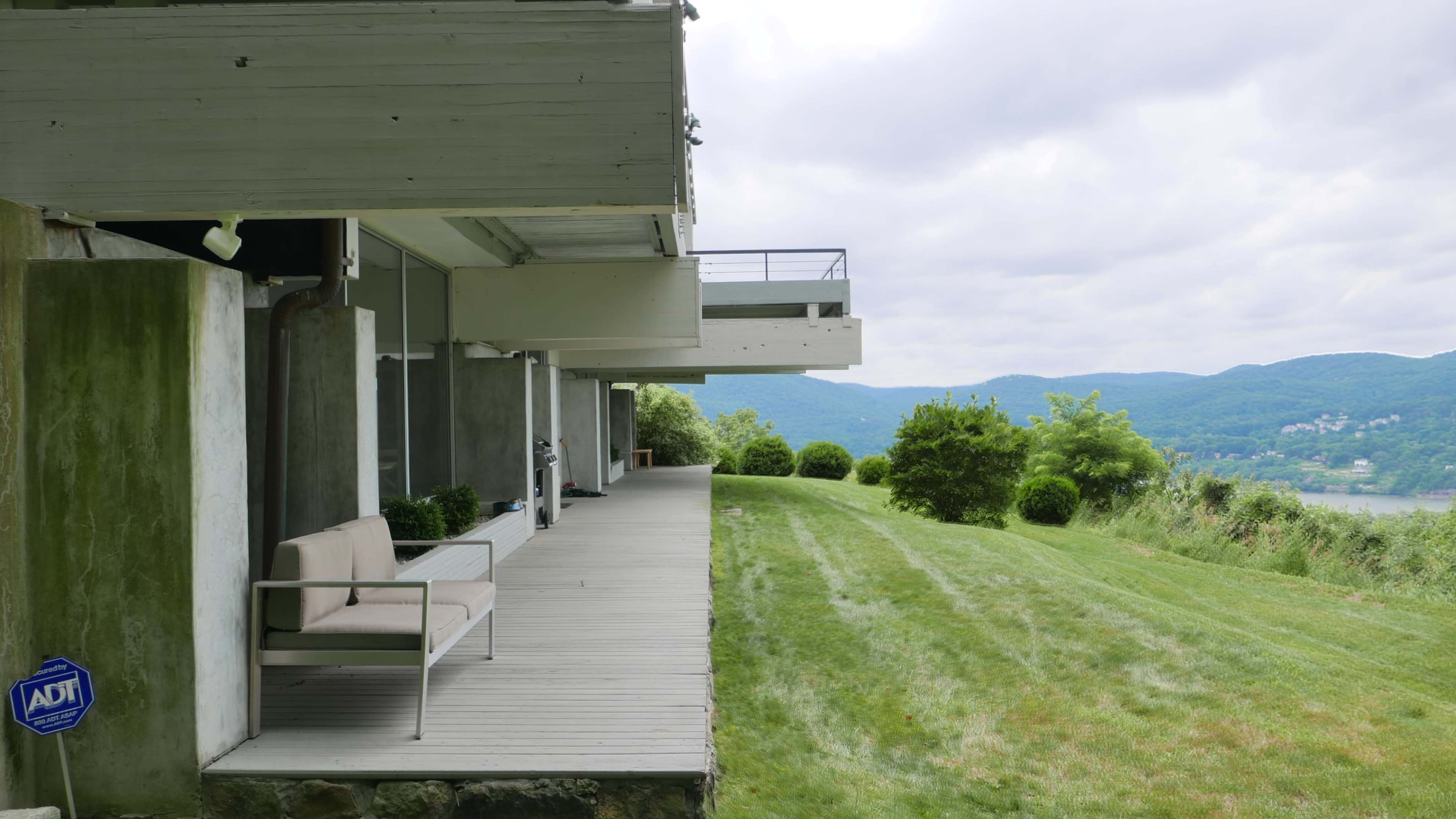 A modern building with a wooden deck and a gray sofa, overlooking a lawn and distant mountains under a cloudy sky.