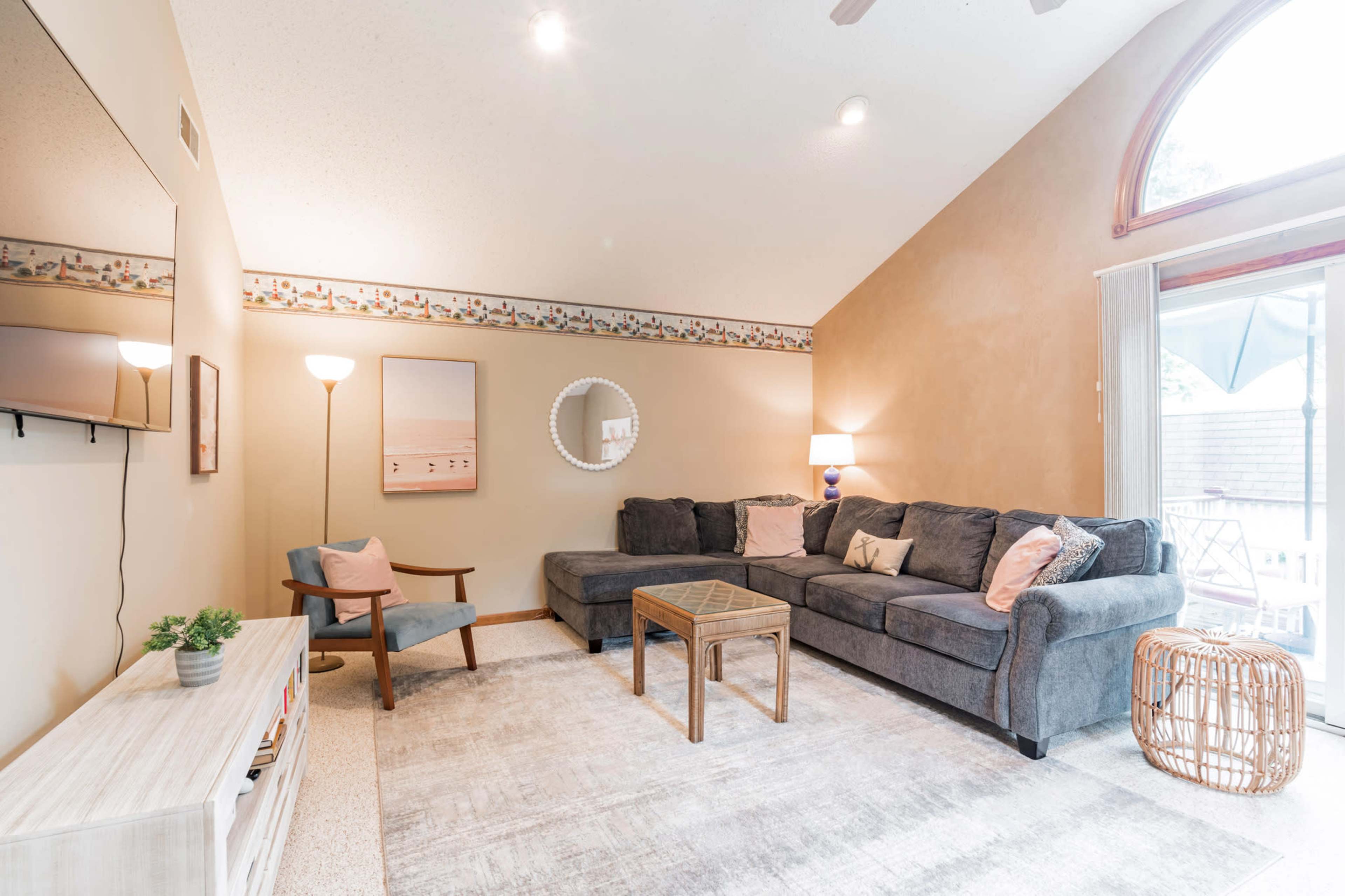 A cozy living room with a grey sectional sofa, a coffee table, a television mounted on the wall, and a large window allowing natural light to enter.