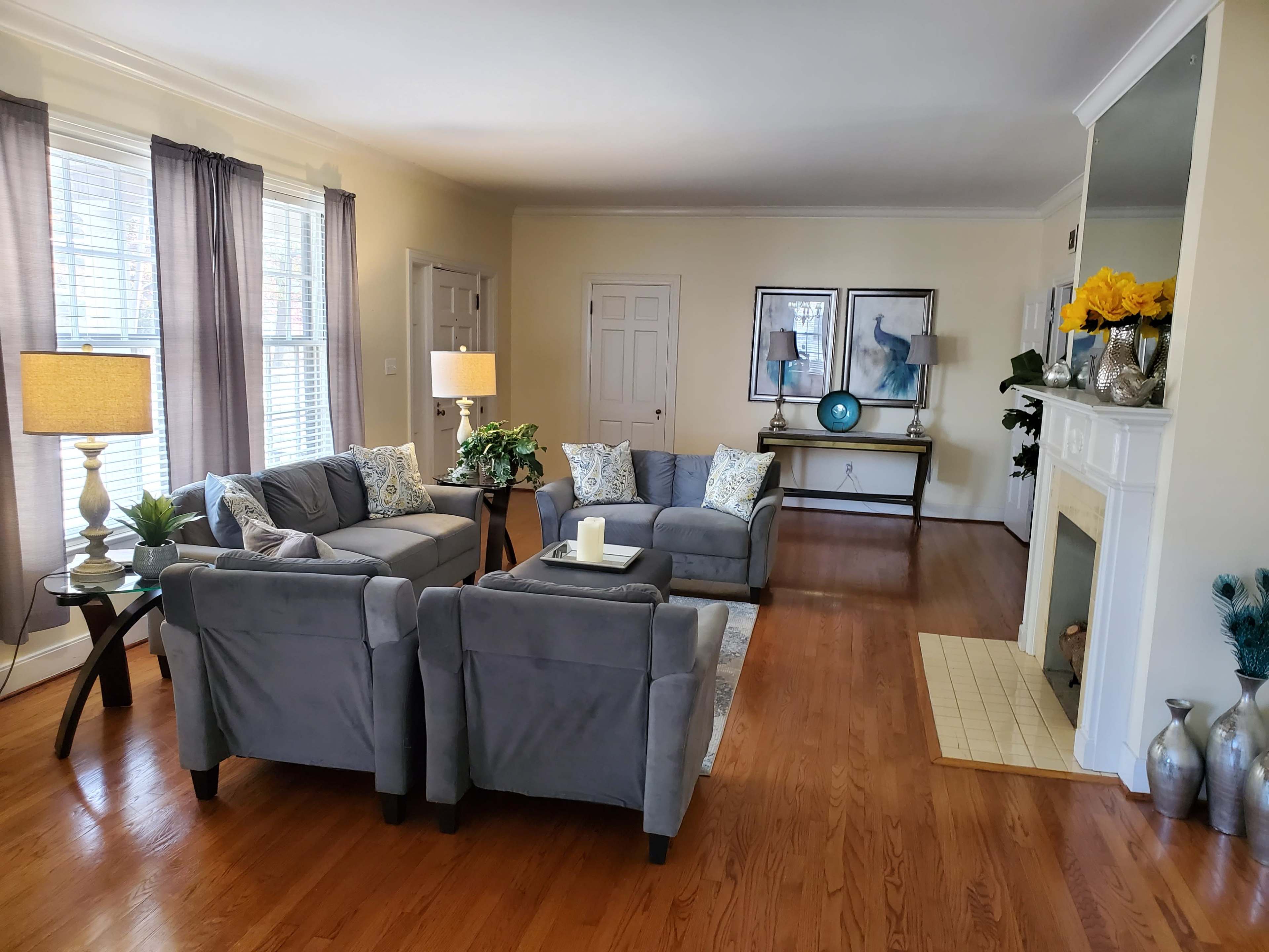A living room furnished with a gray sectional sofa and armchairs, two lamps, and a console table, all set against light-colored walls and wooden flooring.