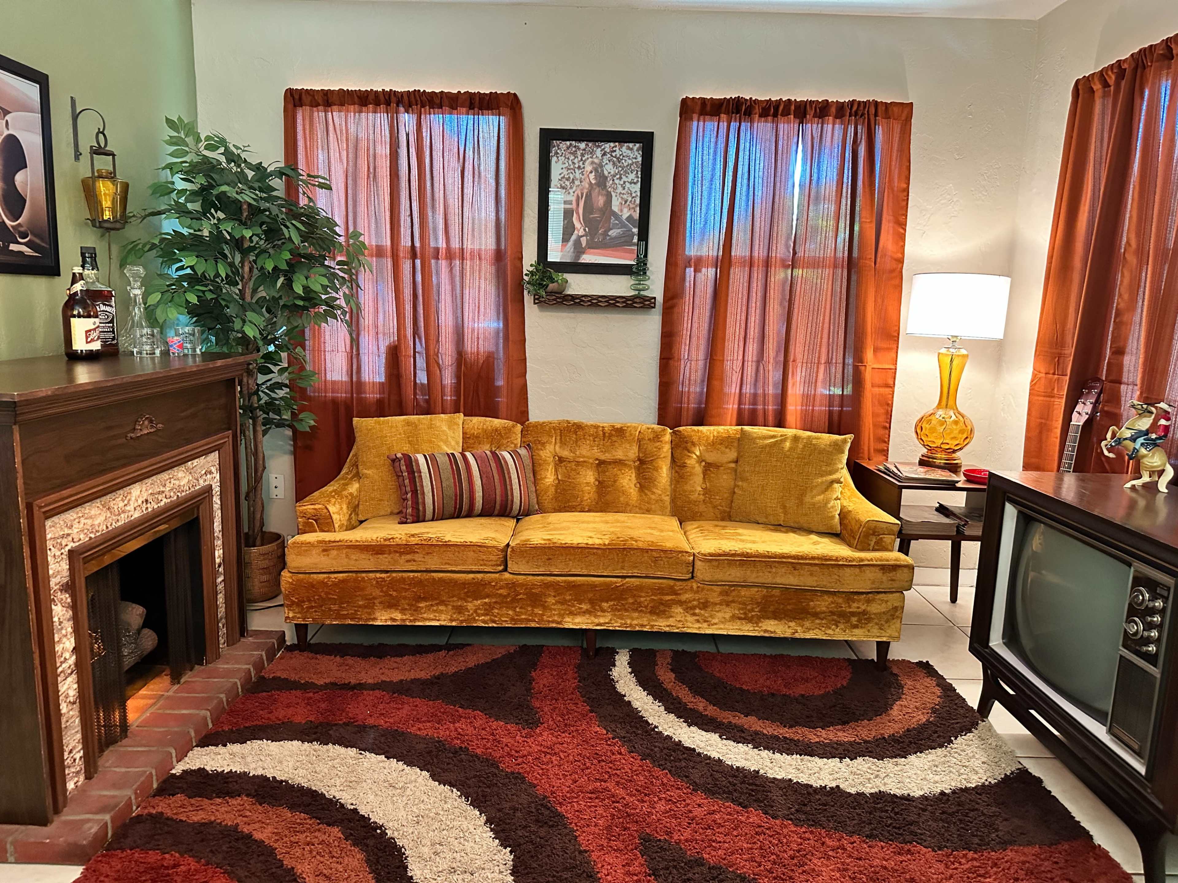 A living room with a yellow velvet couch, a vintage television, a patterned rug, and orange curtains.