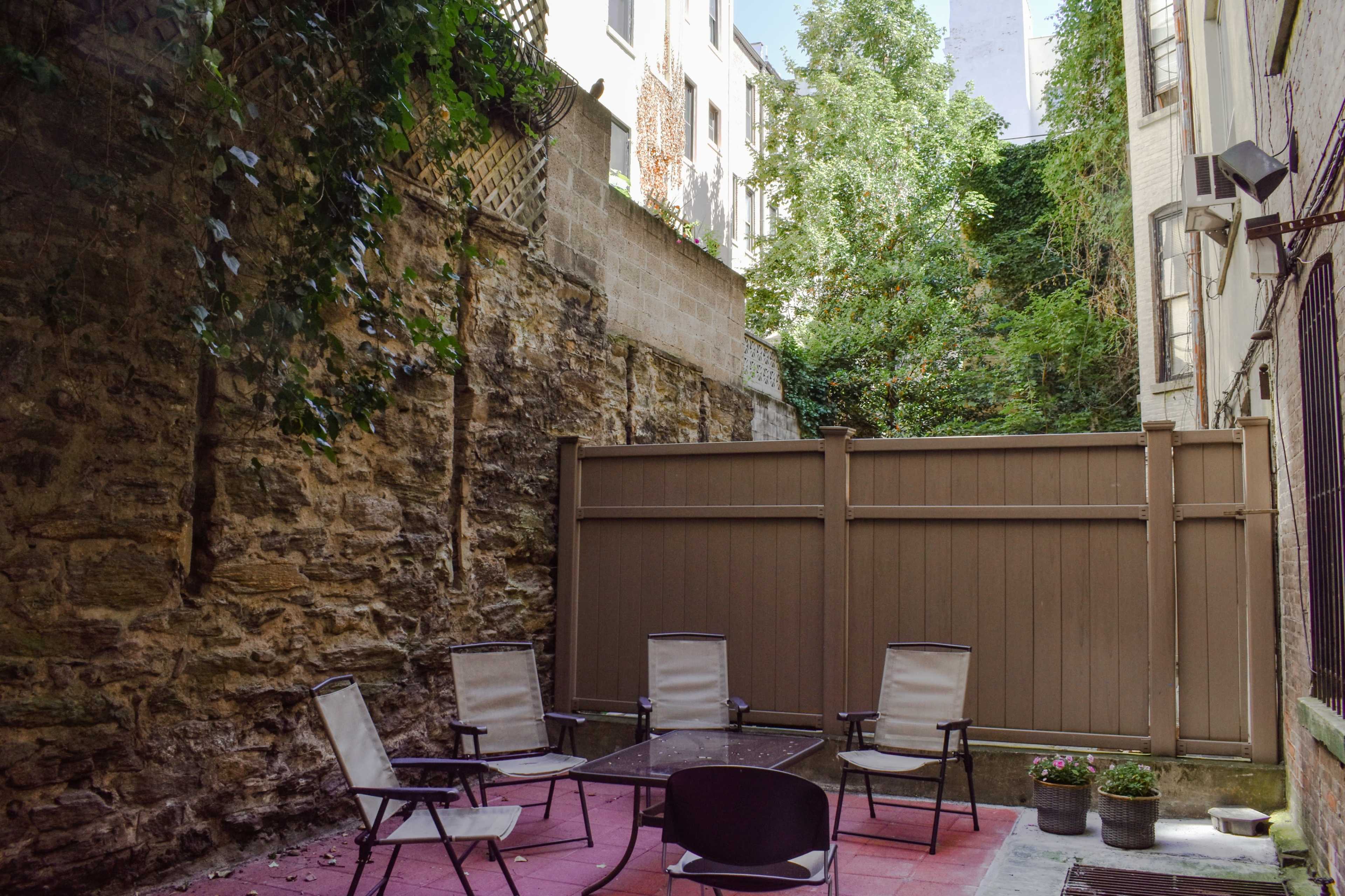 A small patio area features a glass table surrounded by four chairs, with a stone wall and greenery in the background.