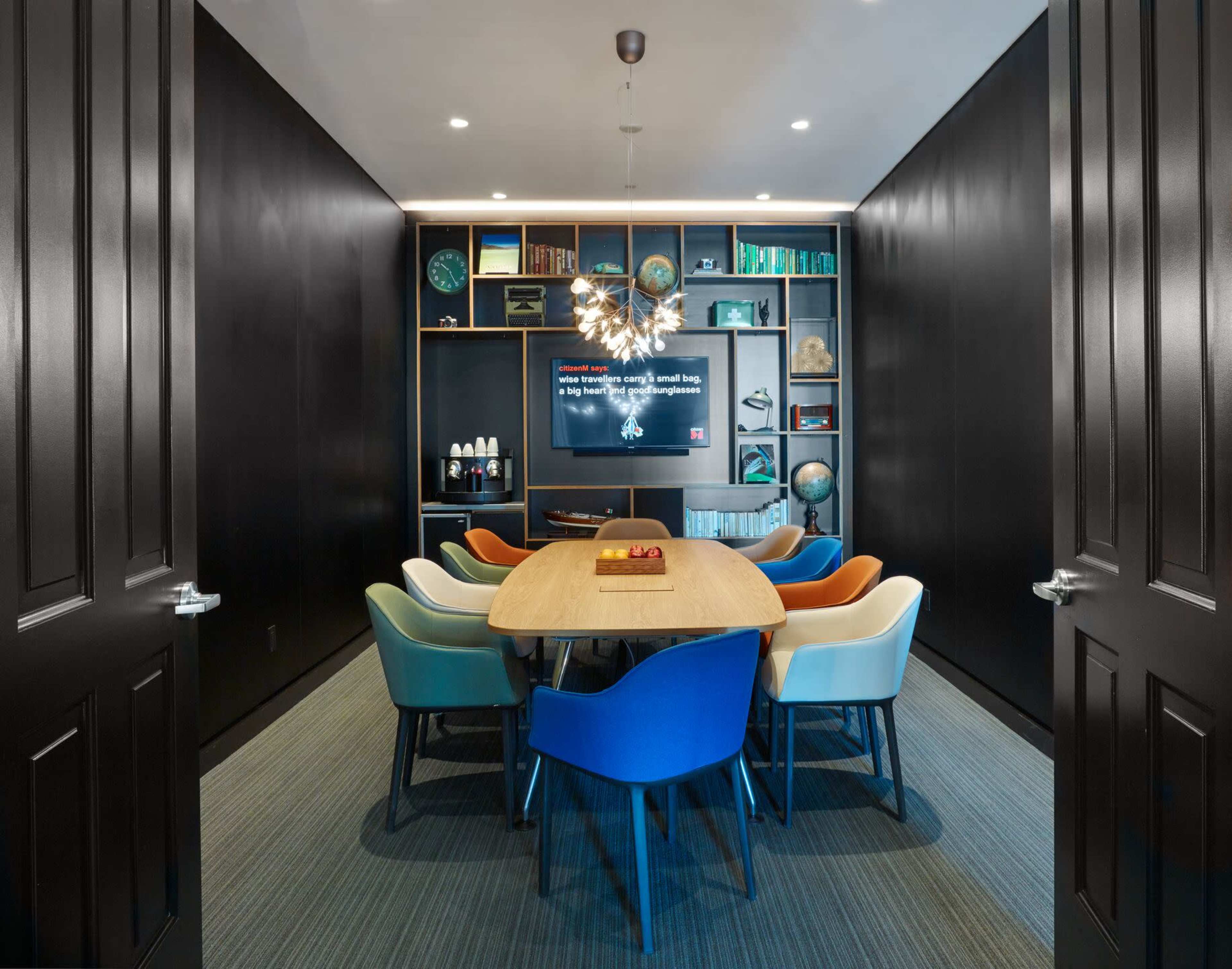 A modern conference room features a large wooden table surrounded by colorful chairs and a television mounted on a black wall, with shelves displaying various items in the background.
