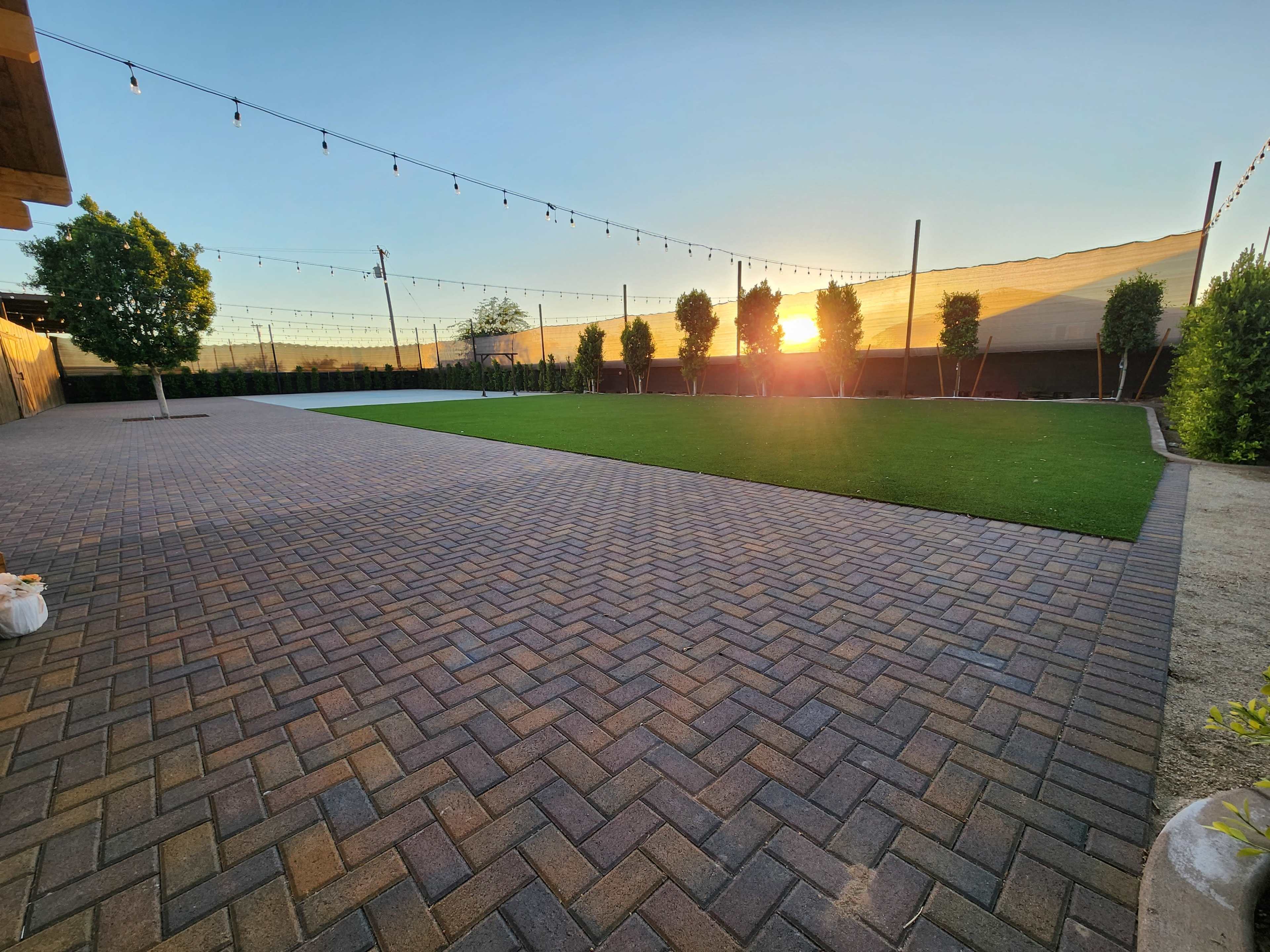 A large, paved outdoor area at sunset, with trees lining the perimeter and string lights overhead.