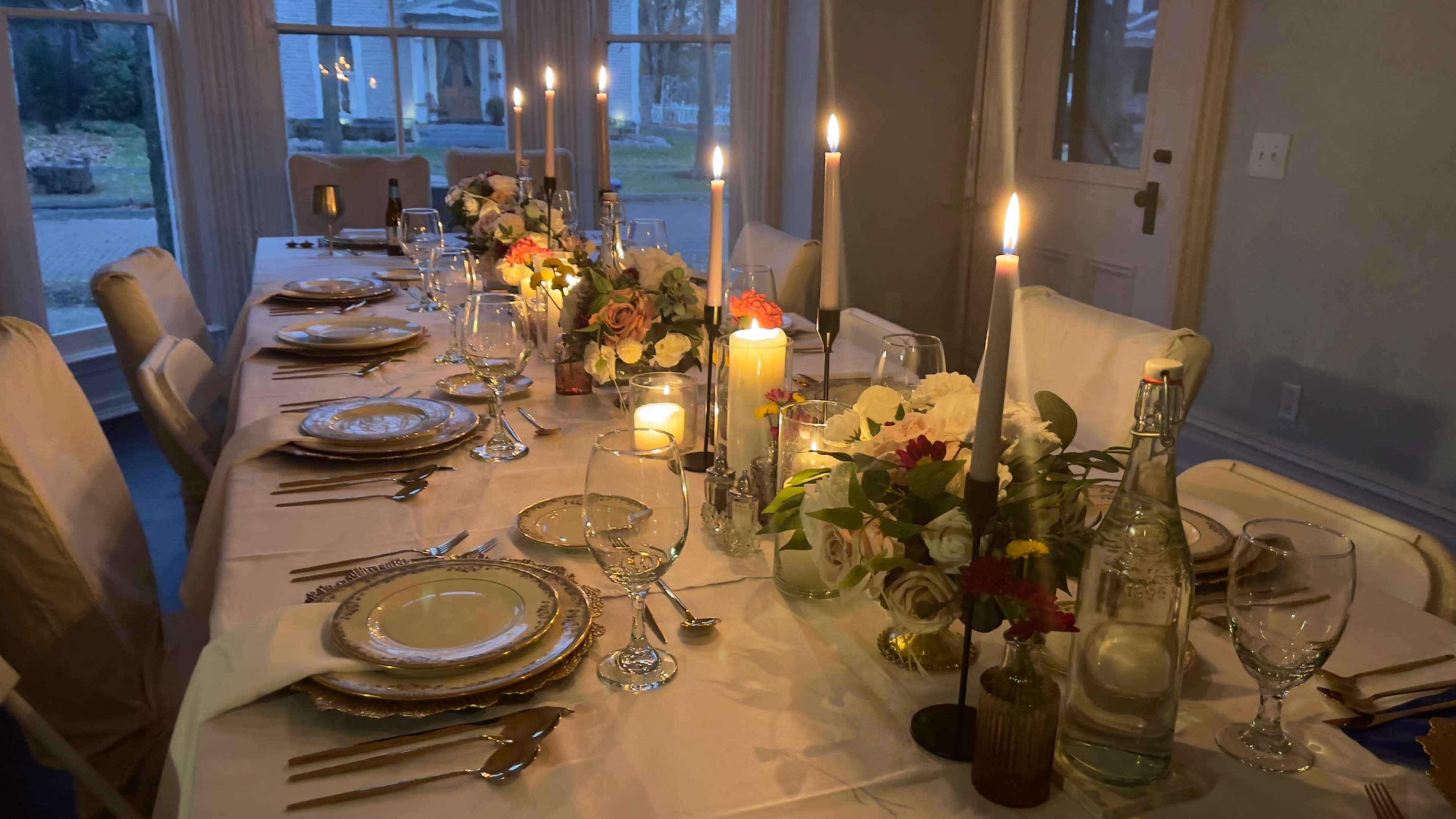 A long dinner table is elegantly set with fine china, glassware, and floral centerpieces, illuminated by candlelight.