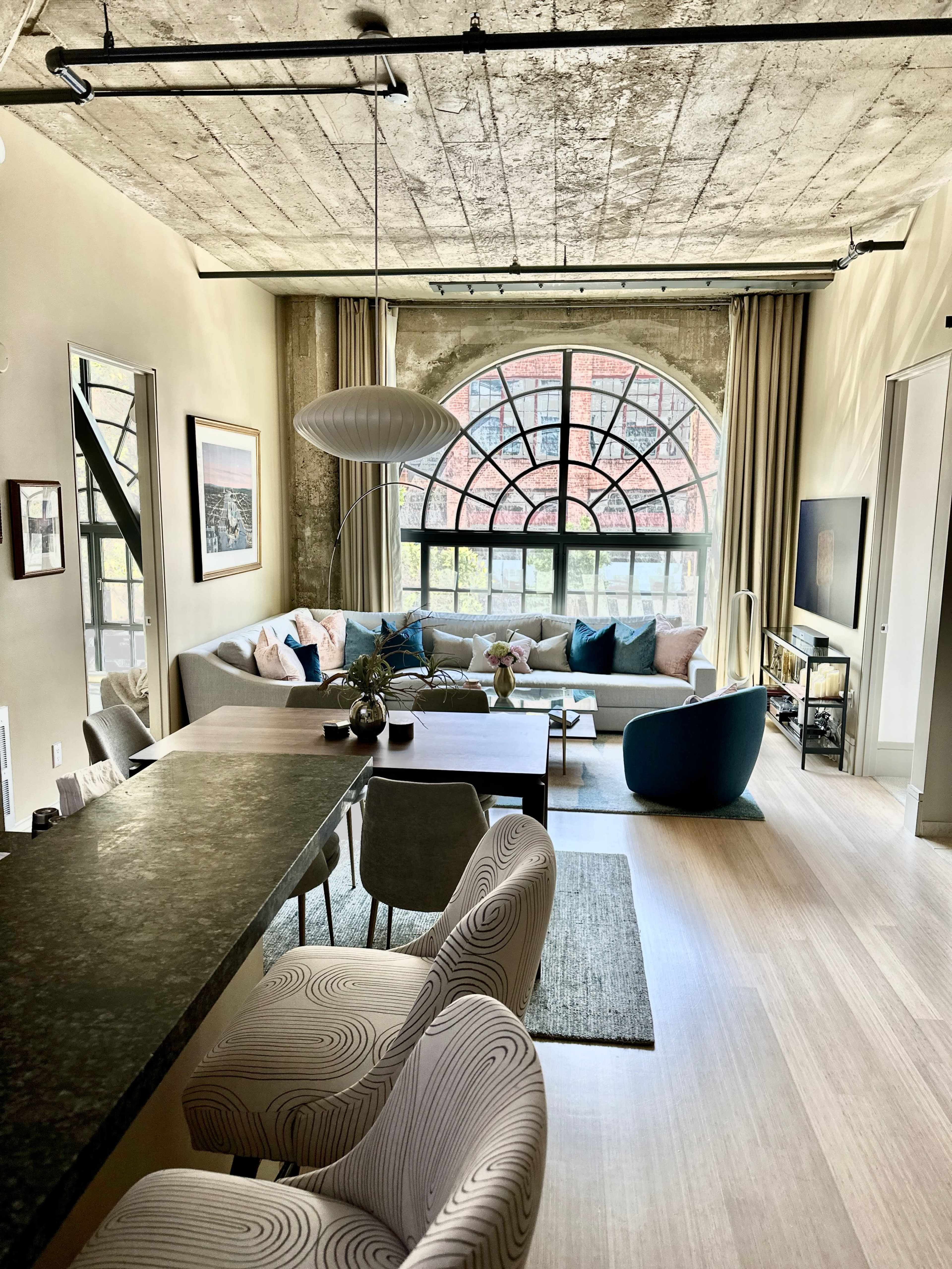 A modern living room with a large arched window, a sectional sofa, a dining area with curved chairs, and a television mounted on the wall.