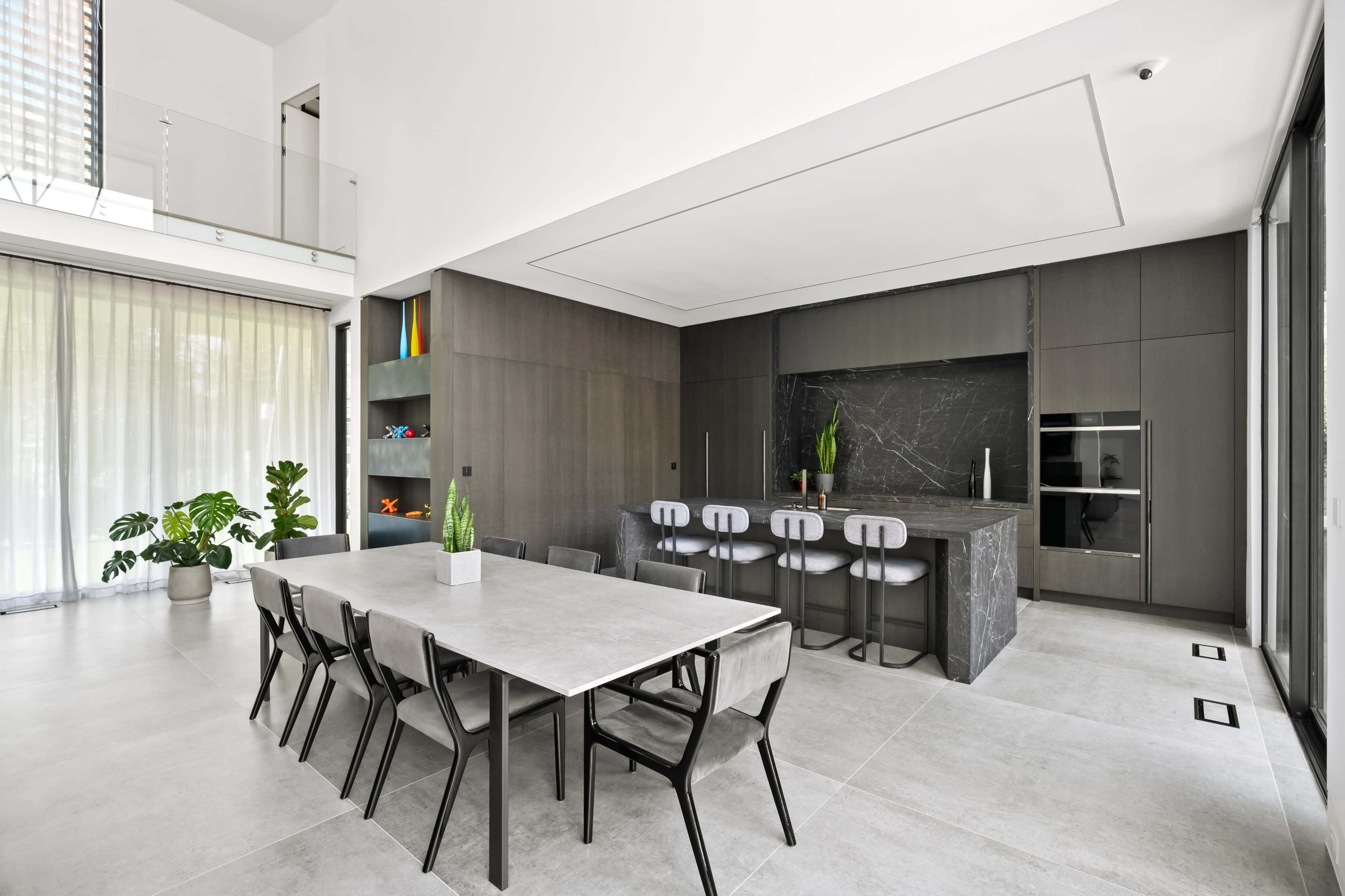 A modern kitchen and dining area features a long concrete table and a dark stone island with seating, surrounded by large windows and indoor plants.