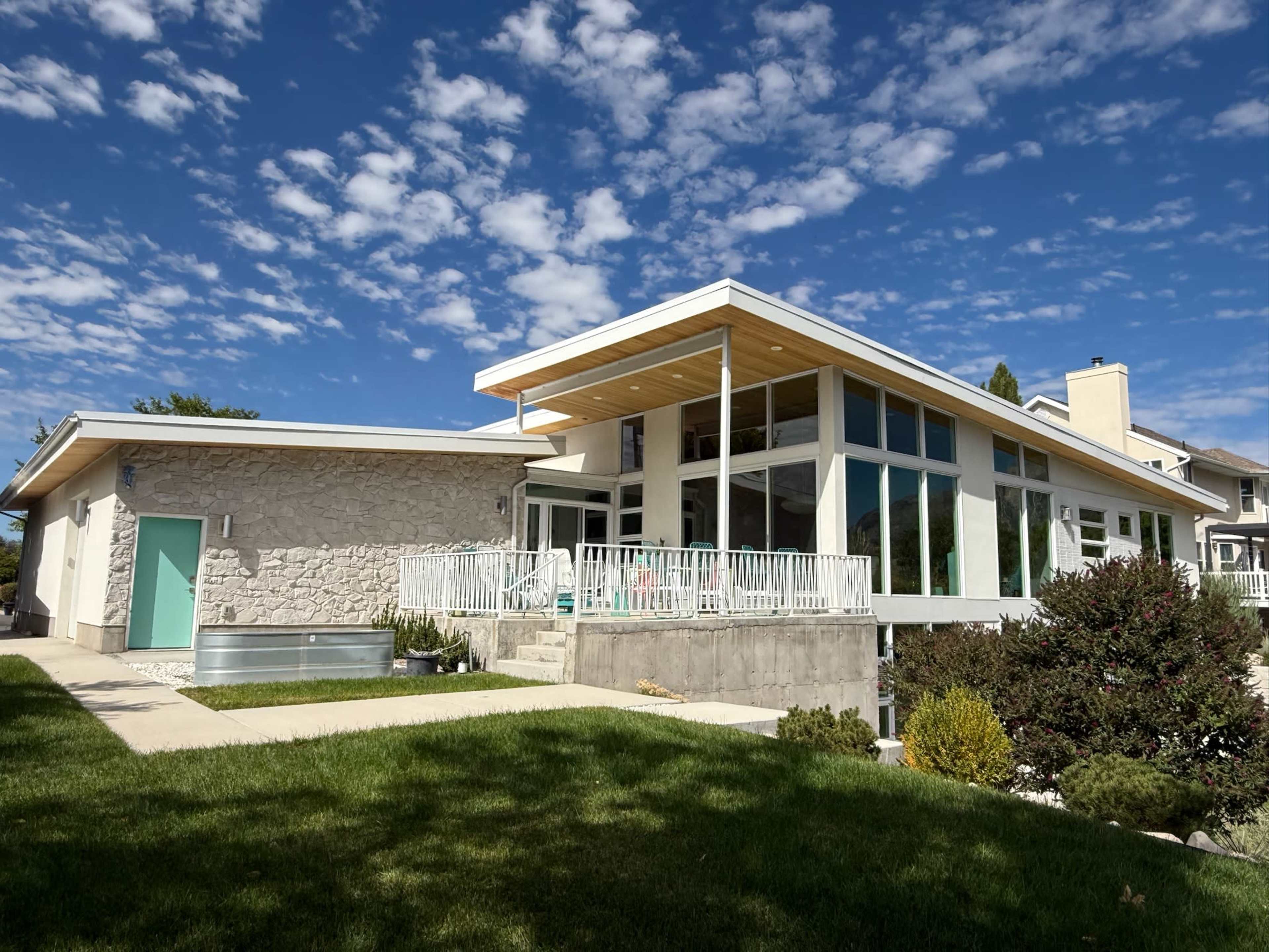 The image shows a modern house with large windows and a sleek design, situated in a landscaped yard under a partly cloudy sky.