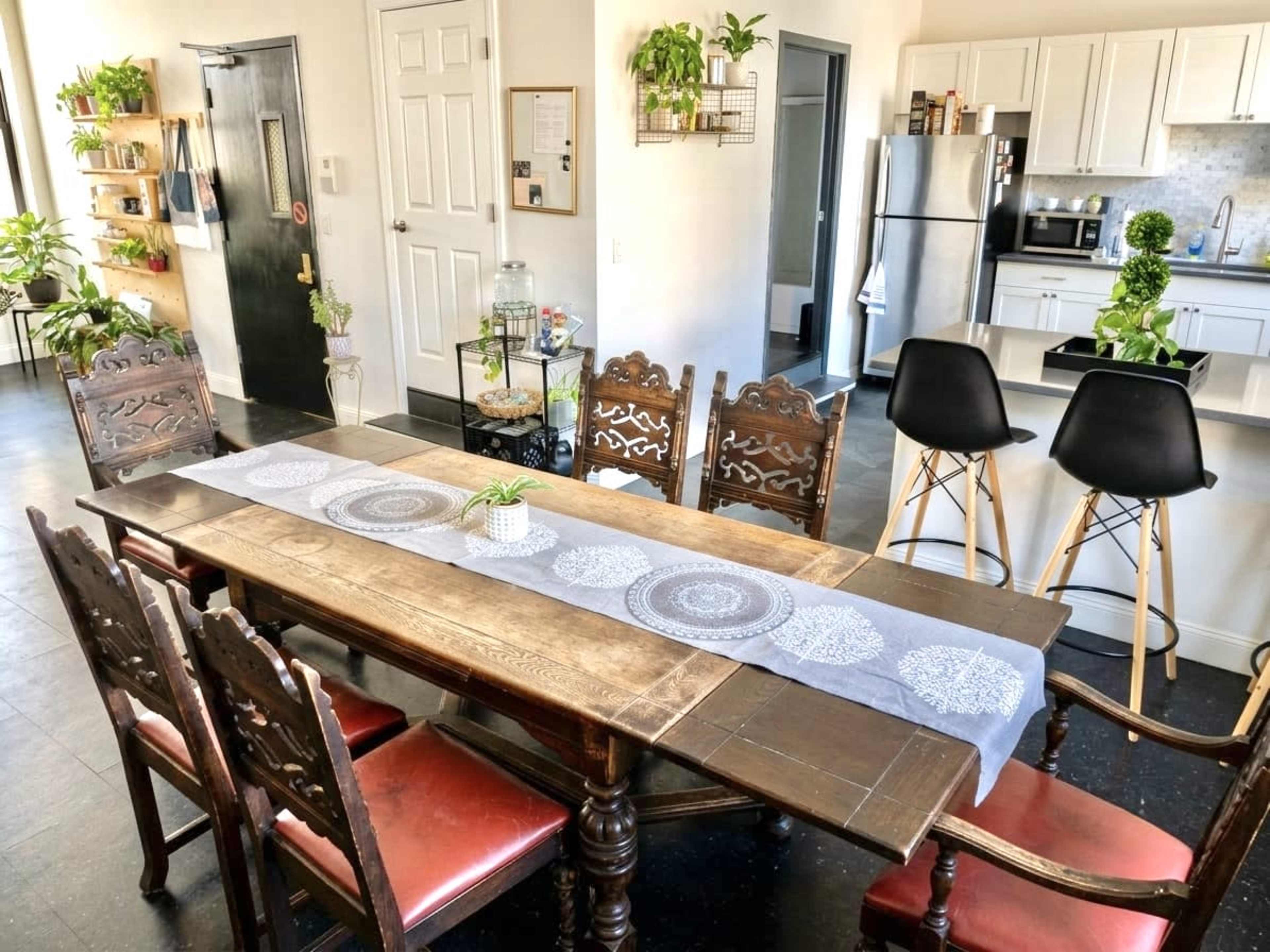 A wooden dining table is set in a bright room with kitchen appliances and decorative plants.