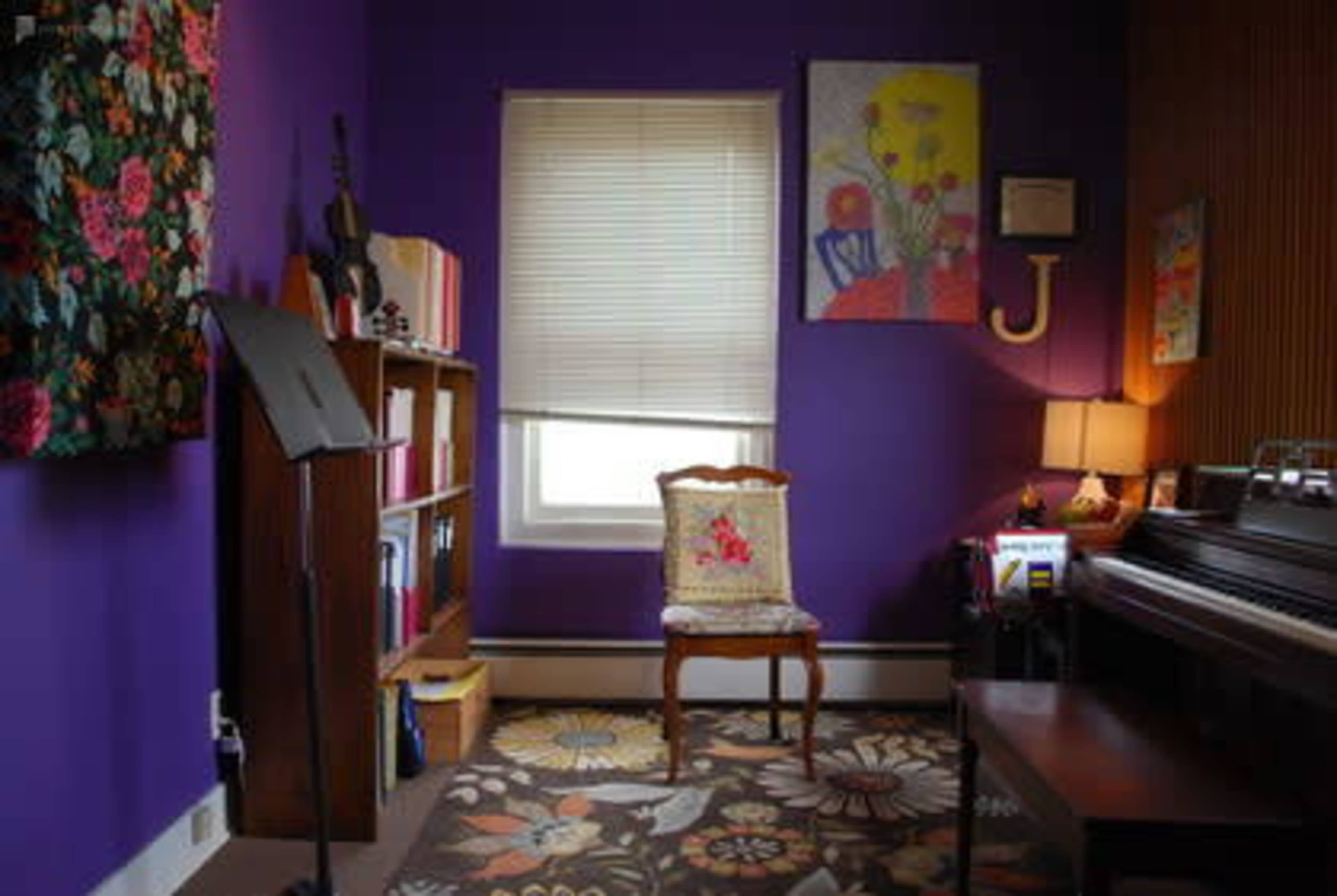 A small room with purple walls, a chair, a bookshelf, and a piano, adorned with floral artwork and a window covered by blinds.
