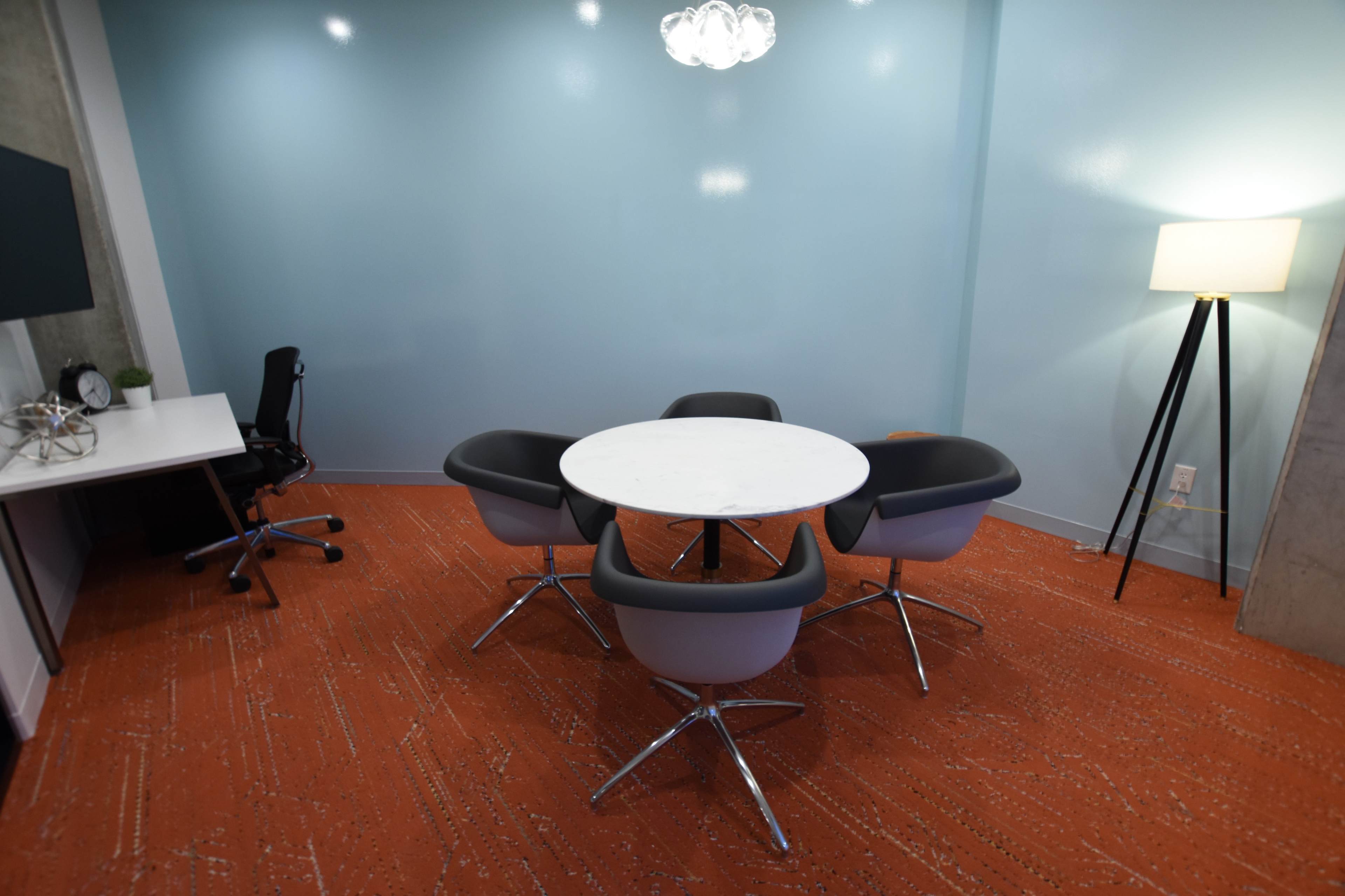 A small conference room features a round table surrounded by four chairs, with a lamp and a desk in the background.
