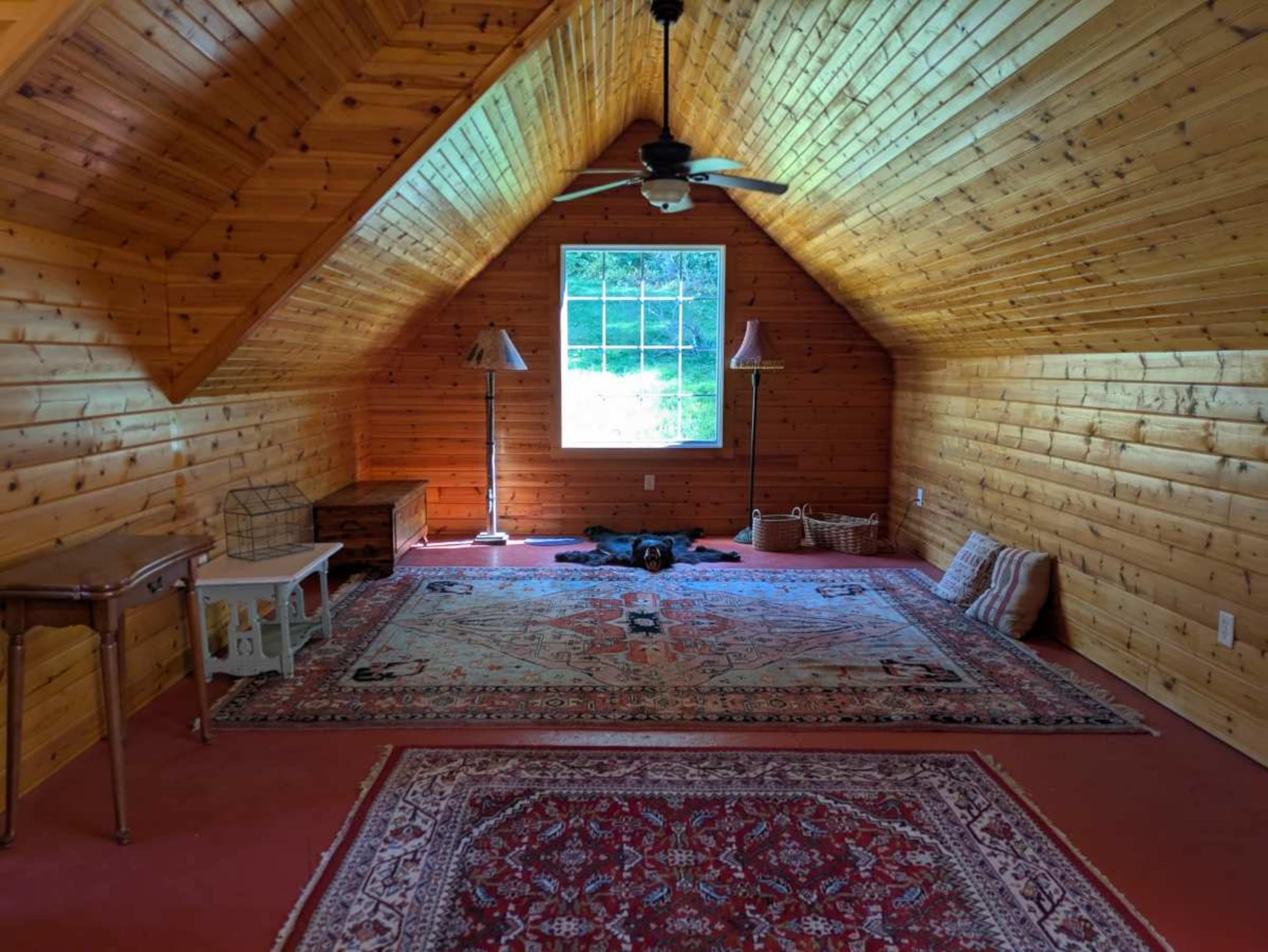 The image shows a spacious attic room with wooden walls and a sloped ceiling, featuring a window, a rug, a table, and a relaxed dog on the floor.