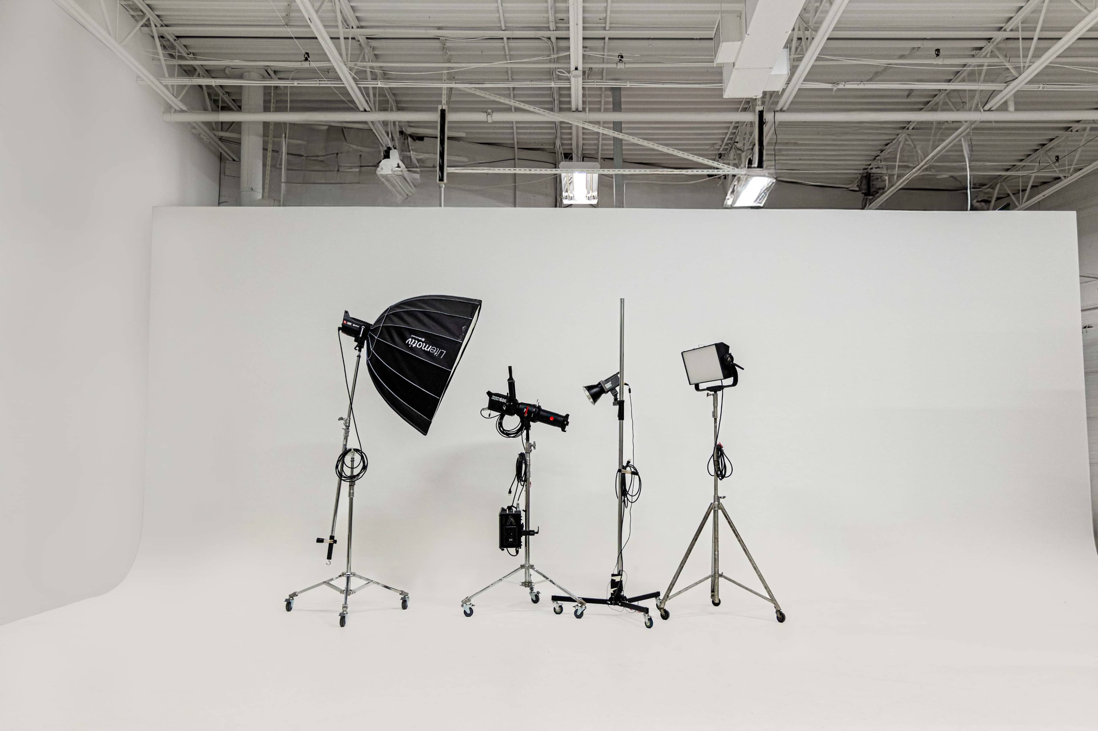 The image shows a photography studio setup with various lighting equipment positioned on stands against a plain white backdrop.