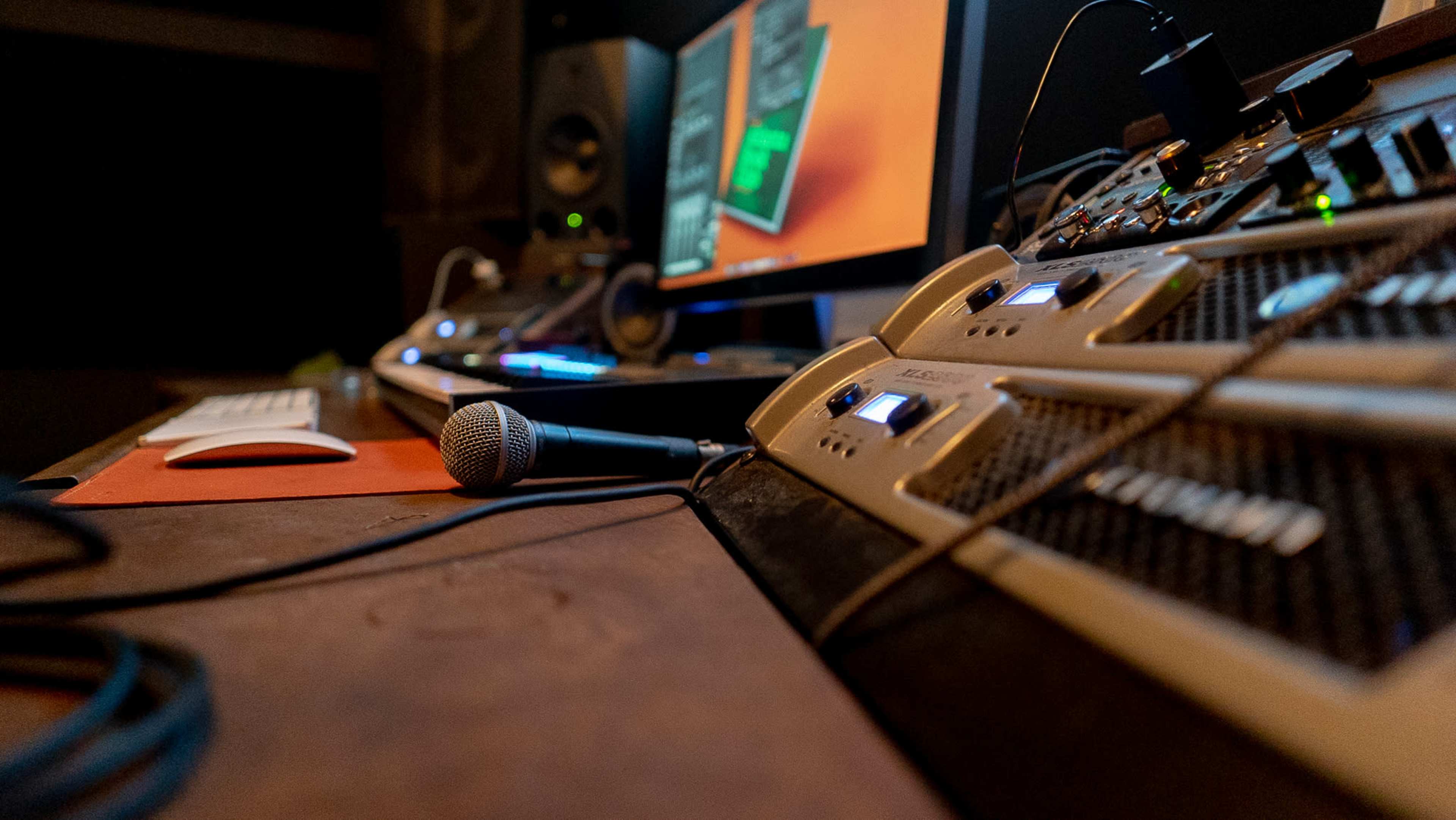 The image shows a close-up view of a sound recording studio with a microphone, computer monitor, and audio equipment on a desk.